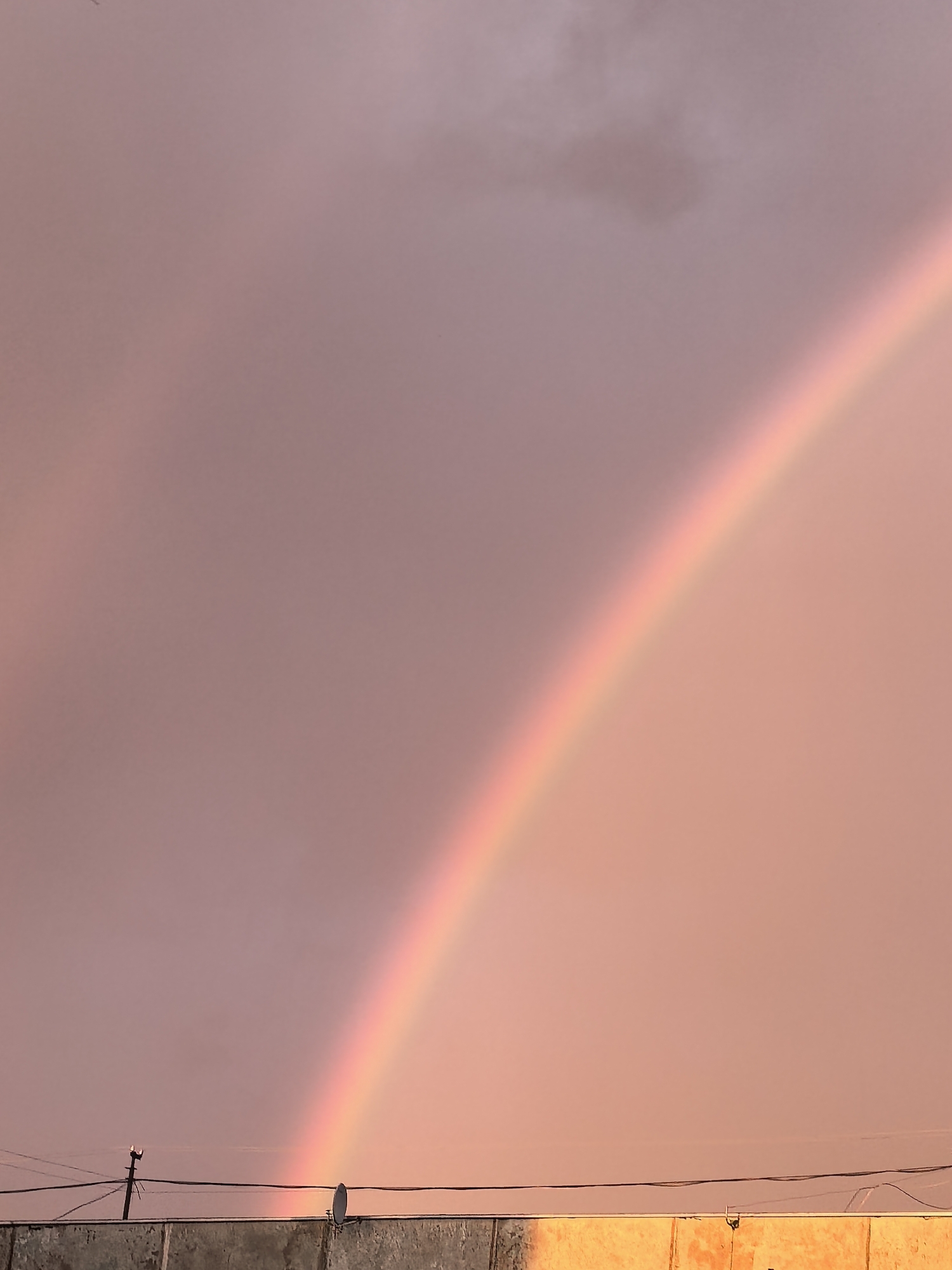 An inverted rainbow arches across a pinkish-mauve sky, its colors muted and blended. The sky is partly cloudy, with some darker patches visible on the left. In the foreground, a flat, gray concrete structure runs horizontally across the bottom of the frame. Power lines are strung tautly above this structure, supported by a dark pole on the left. A small, white satellite dish is affixed to the structure on the right. The overall impression is of a quiet, possibly post-rain, moment in a city or town, with a hint of atmospheric drama from the rainbow. This scene is located in Nerekhta, Russia.