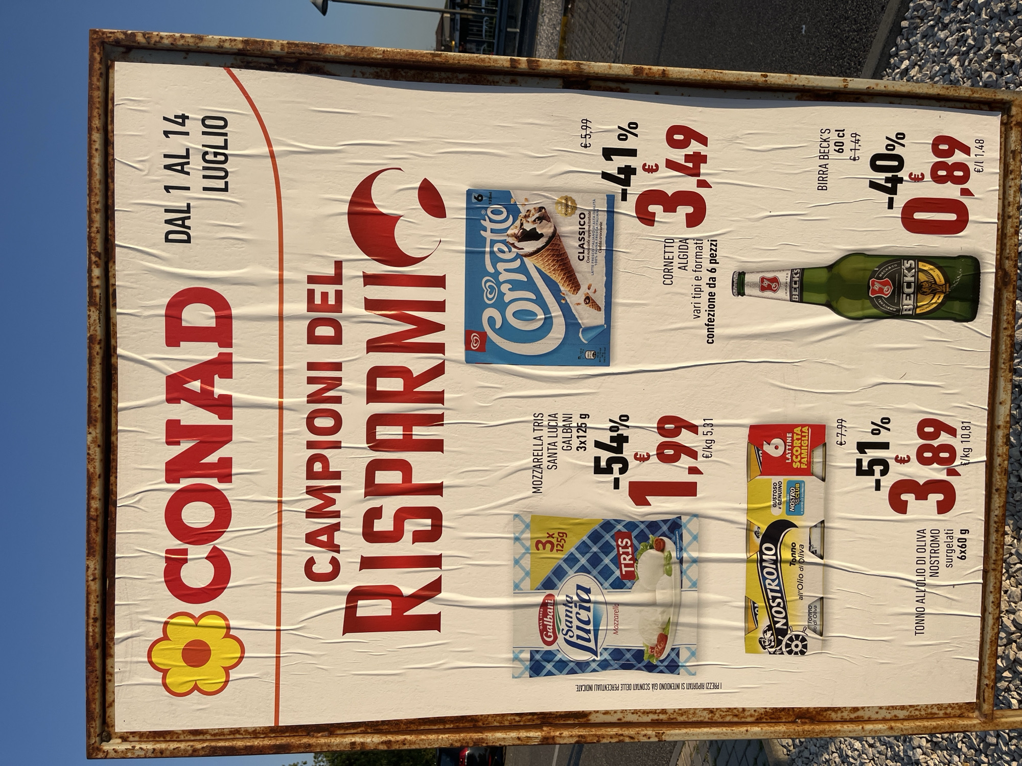 The image shows a large, weathered advertising billboard for CONAD supermarket, located in Castiglione delle Stiviere, Italy. The billboard displays various products and their prices, with a prominent "CAMPIONI DEL RISPARMI" (Champions of Savings) slogan. The products featured include Santa Lucia Mozzarella Tris, Cornetto Algida ice cream, Nostromo canned tuna, and Birra Beck's beer. The billboard is attached to a rusty metal frame and is positioned outdoors, likely in a parking lot or near a road, indicated by the background showing a street lamp and some parked cars. The sky is a clear blue, suggesting daytime. The overall condition of the billboard suggests it has been exposed to the elements for some time, with visible creases and some discoloration on the paper.