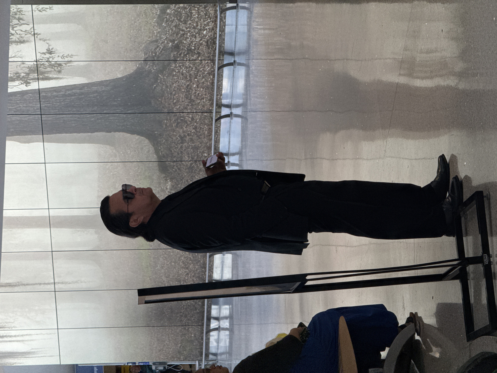 A man wearing a black suit and sunglasses stands in an airport terminal, holding a small white object in his right hand. He is facing a large glass wall, reflecting a tree and outside scenery. Below him, a person is seated, draped with a blue cloth. A black metallic rack partially obscures the view. The location appears to be in an airport terminal, potentially in Kenner, Louisiana, United States of America. The man appears to be observing the reflection on the window.