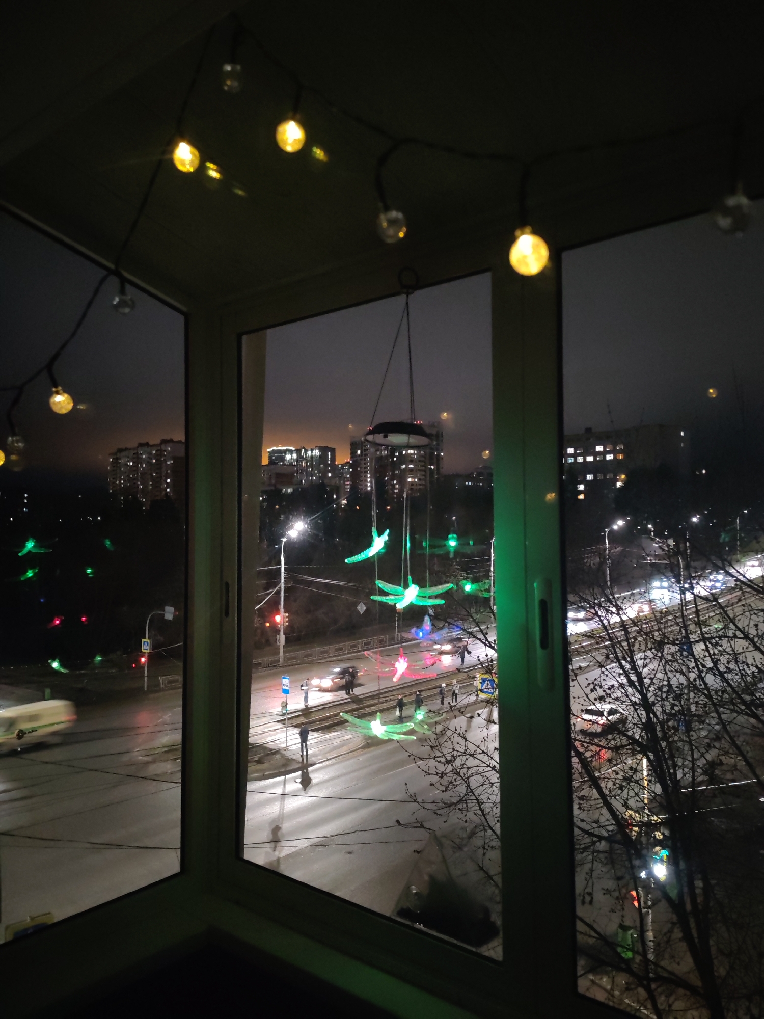 A nighttime view from an indoor window showing a street scene in Izhevsk, Russia. The scene includes a road with cars and pedestrians crossing at crosswalks. Trees are visible on the right side of the frame. Overhead, fairy lights hang indoors. Reflective surfaces of wet roads and building windows enhance the lighting effects. A decorative wind chime is visible, hanging in front of the window. The urban environment comprises buildings with lit windows in the background.