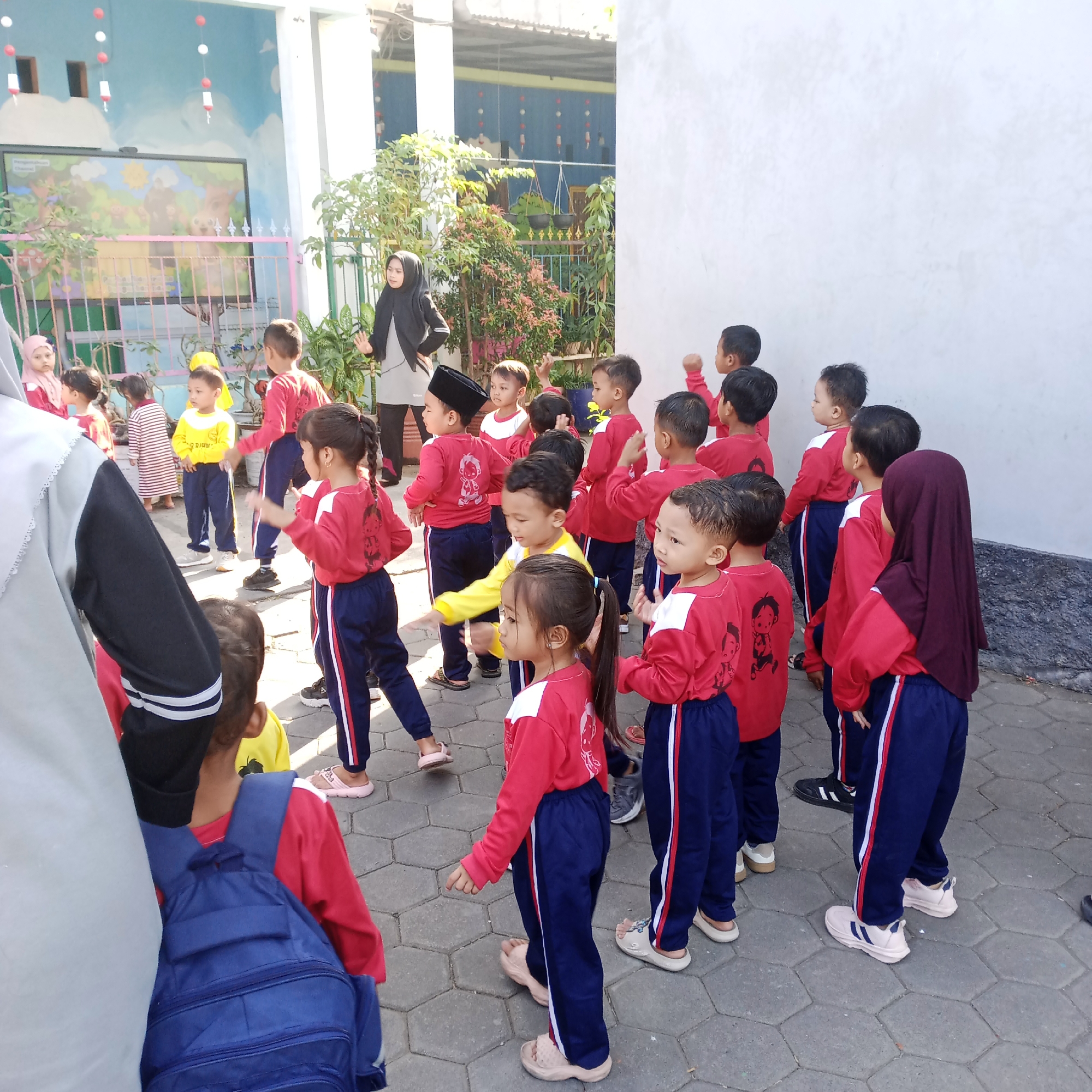 In Rejosari, Indonesia, a group of young children, likely in a school setting, are gathered outdoors. They are dressed in matching red shirts and blue pants with red and white stripes. A female adult, wearing a black hijab and a gray top, stands to the left, appearing to be addressing or instructing the children. Several children are looking in different directions, some with hands raised. The environment includes a white building with a painted mural on its exterior wall, a green fence, and lush green plants. The ground is paved with hexagonal gray tiles. The overall scene suggests an organized activity or lesson for the children.