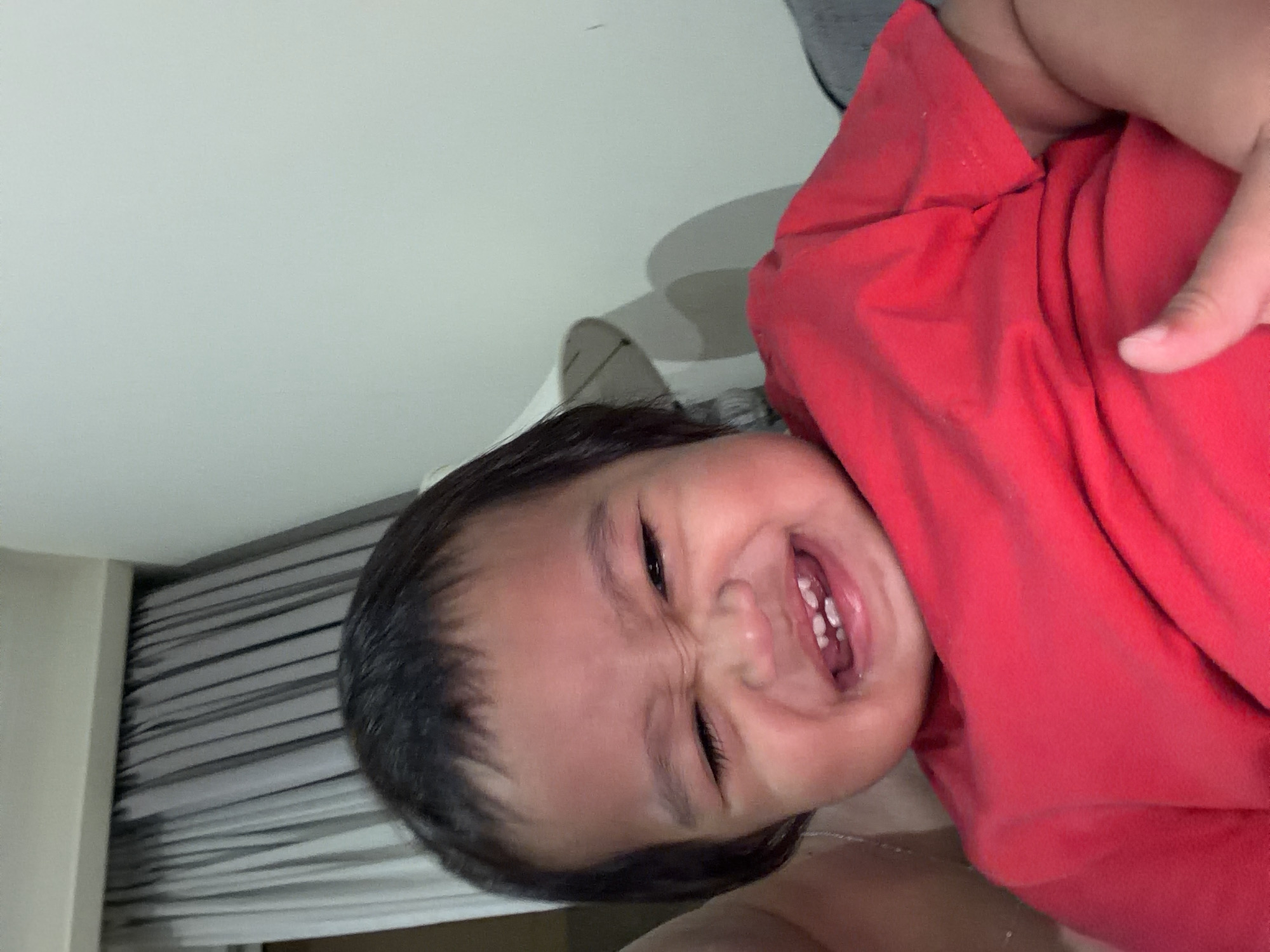 The image shows a close-up of a young child wearing a red shirt, grimacing with their eyes squeezed shut and teeth showing. The child is being held, with a thumb visible in the foreground. The background consists of a wall and curtains. There is no information in the image to suggest a location in Central Jakarta, Indonesia.