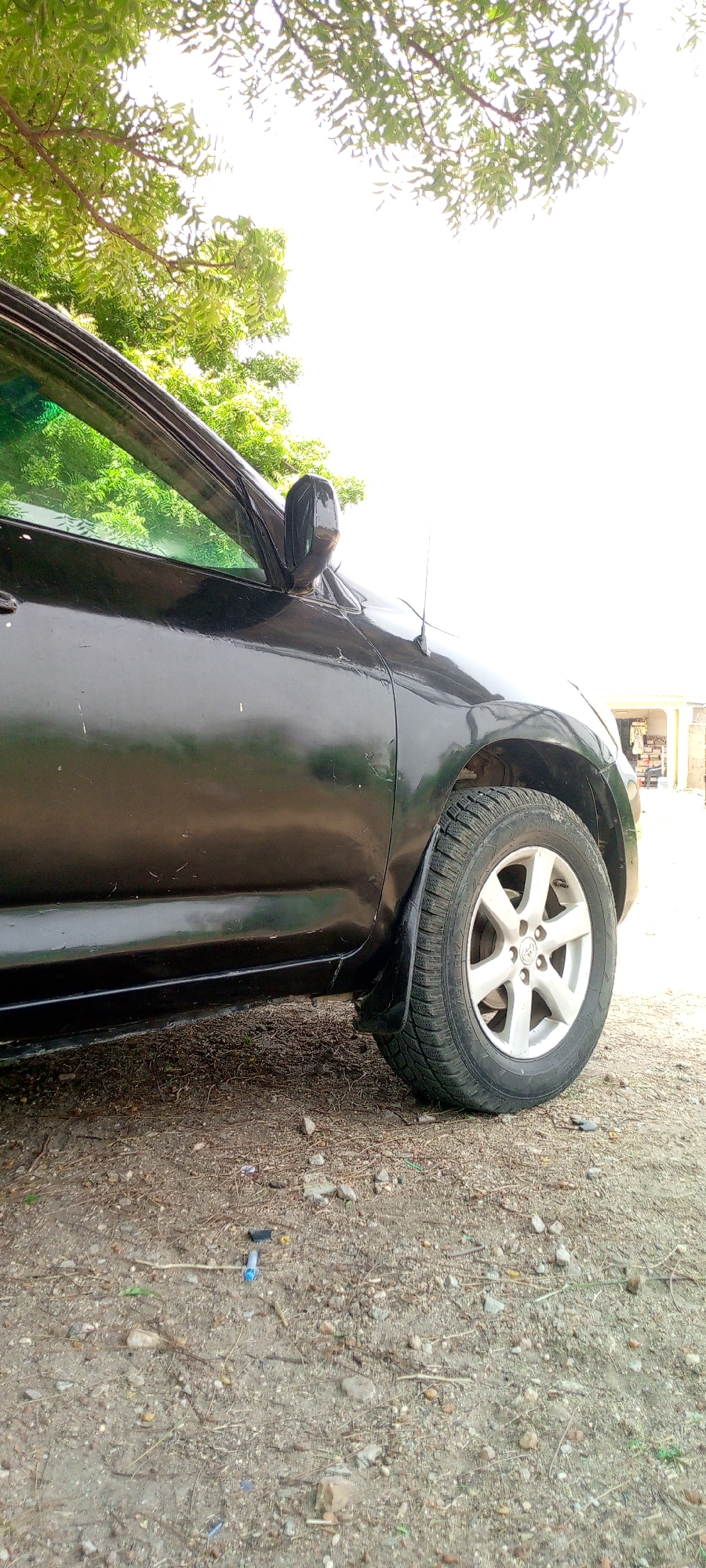 A black car, likely a Toyota RAV4, is parked on a dirt and gravel surface in what appears to be Adankolo, Nigeria. The image focuses on the front driver's side of the vehicle, showing the front fender, tire, and side mirror. The car's paint shows signs of wear. A tree with green foliage overhangs the car. The background is bright, possibly indicating sunlight. Debris, including small pieces of trash, is scattered on the ground.