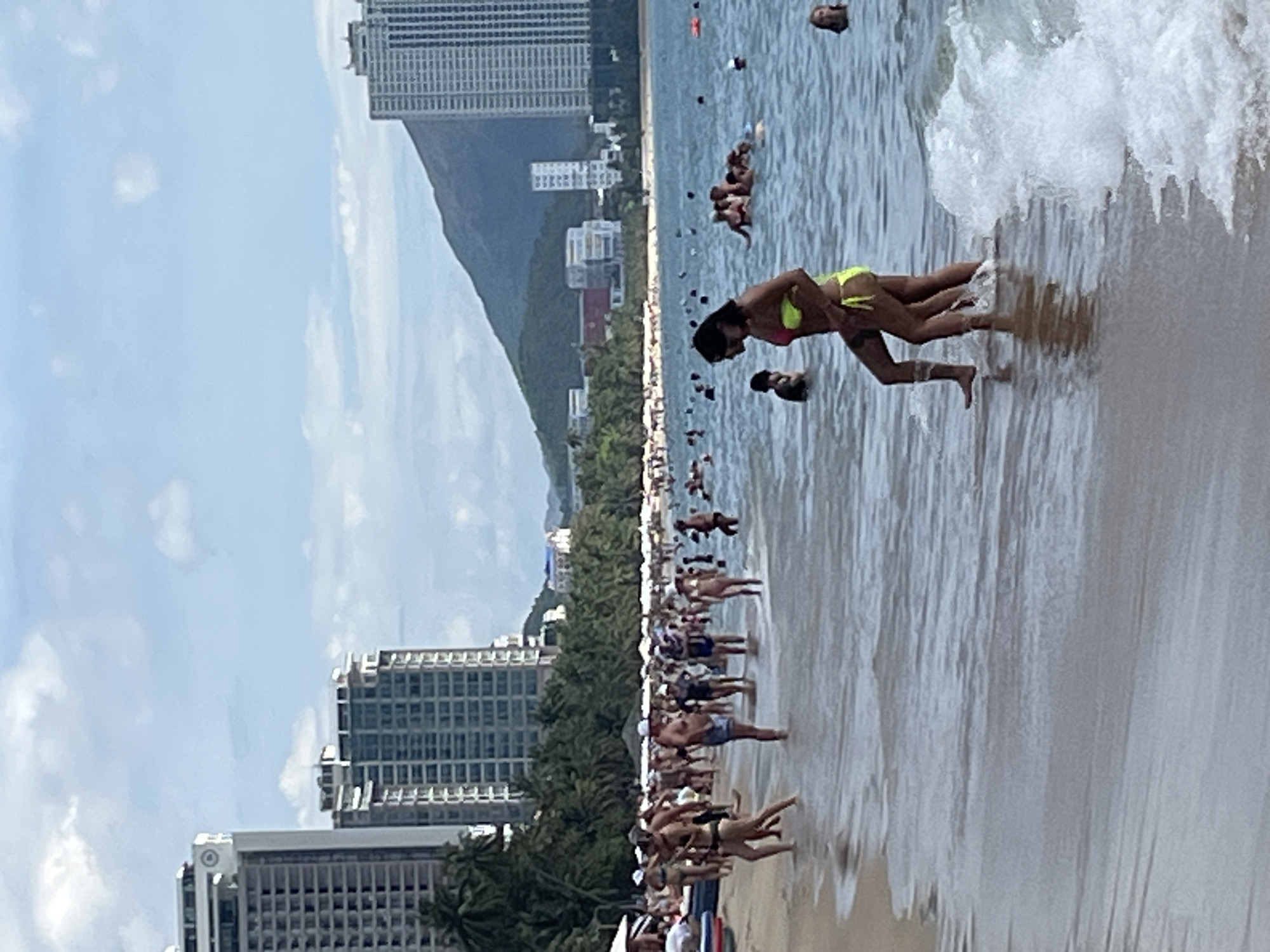 In Nha Trang, Vietnam, a crowded beach scene unfolds under a partially cloudy sky. High-rise buildings, characteristic of the city's coastal development, dominate the background, with palm trees and lower structures nestled between them and the mountains. The foreground is occupied by the ocean, with waves gently breaking on the shore. A diverse group of people, dressed in swimwear, are engaged in various beach activities. Some are wading in the shallow water, others are walking along the sandy shore, and a significant number are enjoying the sea, with a few individuals in the water further out. The overall atmosphere suggests a popular tourist destination bustling with activity on a warm day.