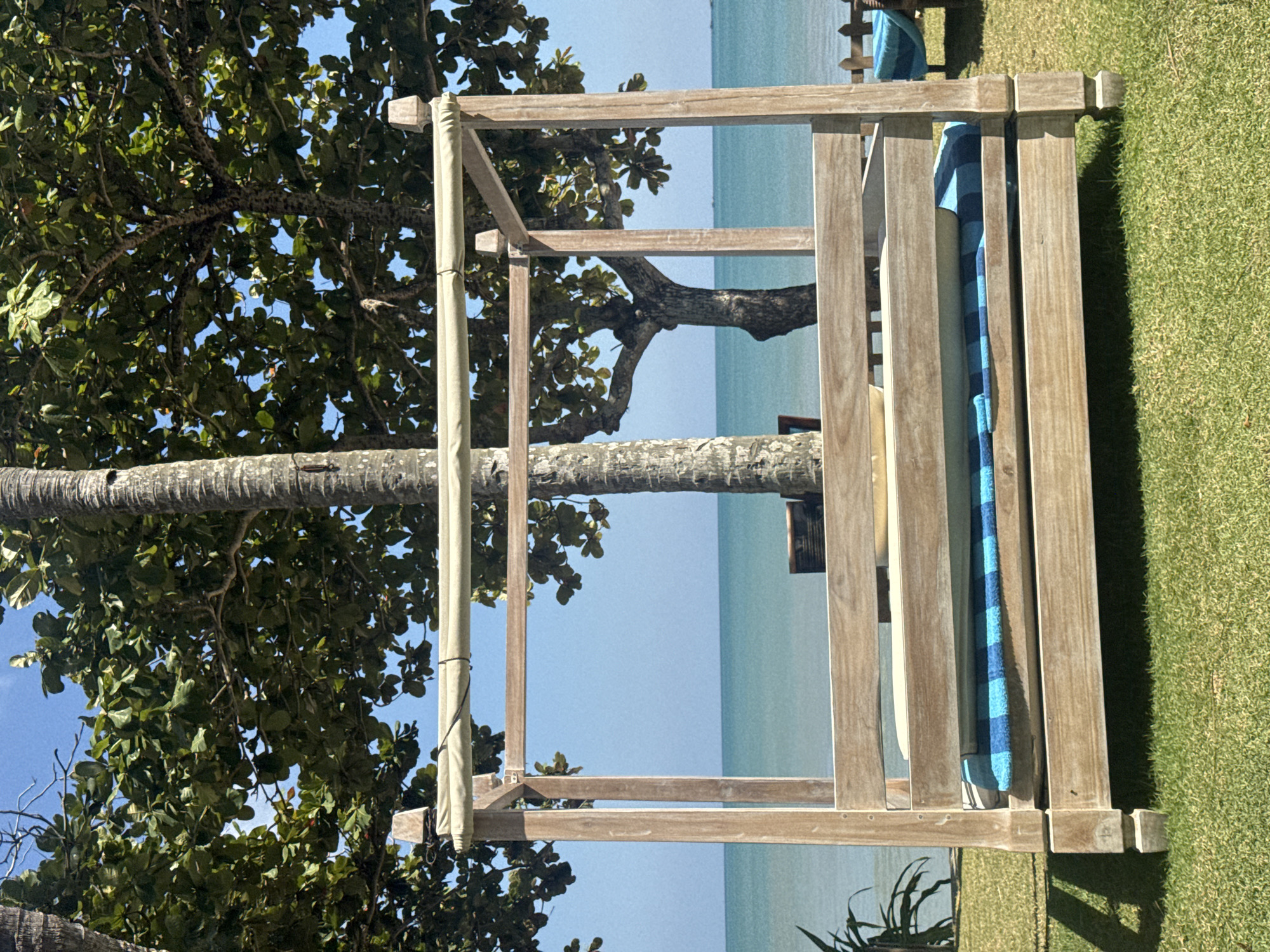 In Kuta Selatan, Indonesia, a wooden four-poster daybed with a blue and white striped cushion sits on a manicured green lawn. The daybed is positioned in front of a tranquil turquoise ocean under a clear blue sky. Lush green trees frame the scene, with the trunk of a palm tree visible to the left and a larger tree with dark bark to the right. The ocean extends to the horizon, suggesting a peaceful coastal setting.
