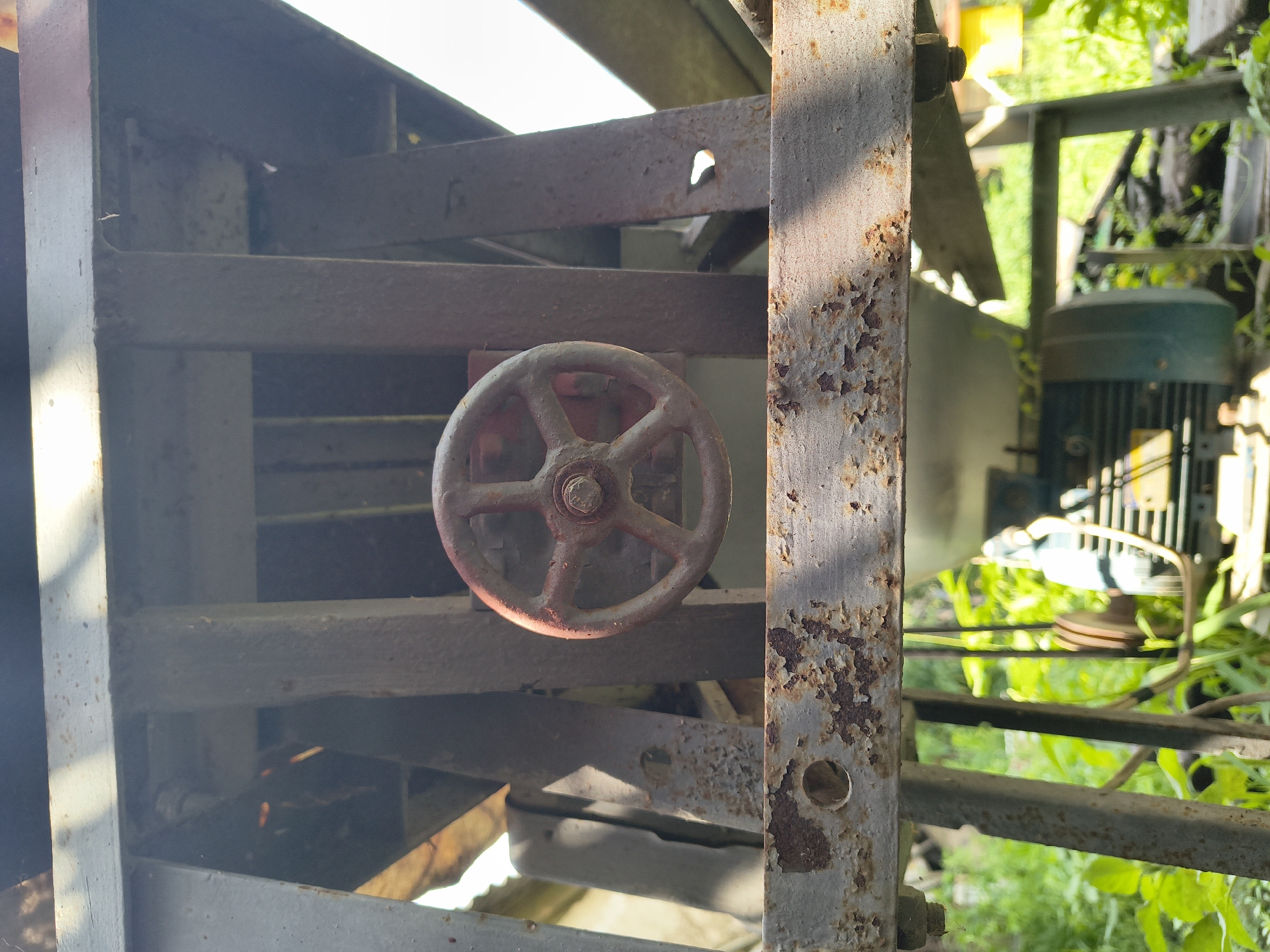 The image shows a close-up of a rusty red handwheel on a metal structure, likely part of an old machine or industrial equipment. The handwheel has five spokes and a central bolt. The metal structure itself is weathered and shows signs of rust and peeling paint. Behind the handwheel, the background is blurred but suggests an outdoor environment with greenery and sunlight filtering through. Further in the background, a motor with a pulley system is partially visible. The overall impression is one of disuse and decay. The image does not contain any people or provide any specific clues about its location in Mikhaylovka, Russia, or any other specific city.