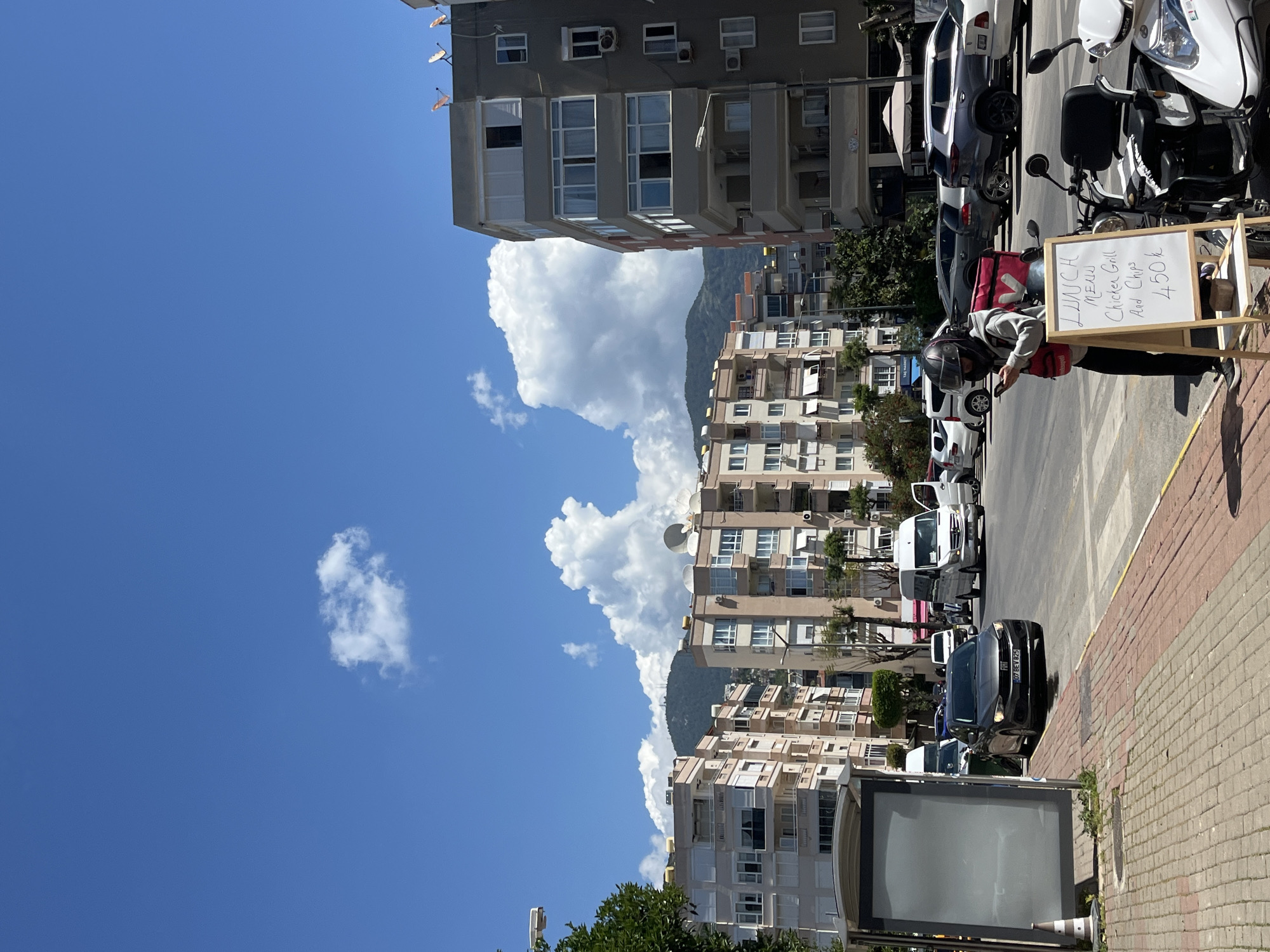 The image depicts a street scene in Alanya, Turkey, with a clear blue sky punctuated by cumulus clouds, positioned above a mountainous backdrop. A row of apartment buildings lines one side of the street, various parked cars are visible alongside the opposite side of the street, and a man wearing a helmet and red backpack is standing next to a small easel advertising a lunch menu. The menu board reads "Lunch Menu: Chicken Grill and Chips 450k." Several parked scooters are nearby. The foreground consists of a brick-paved sidewalk.
