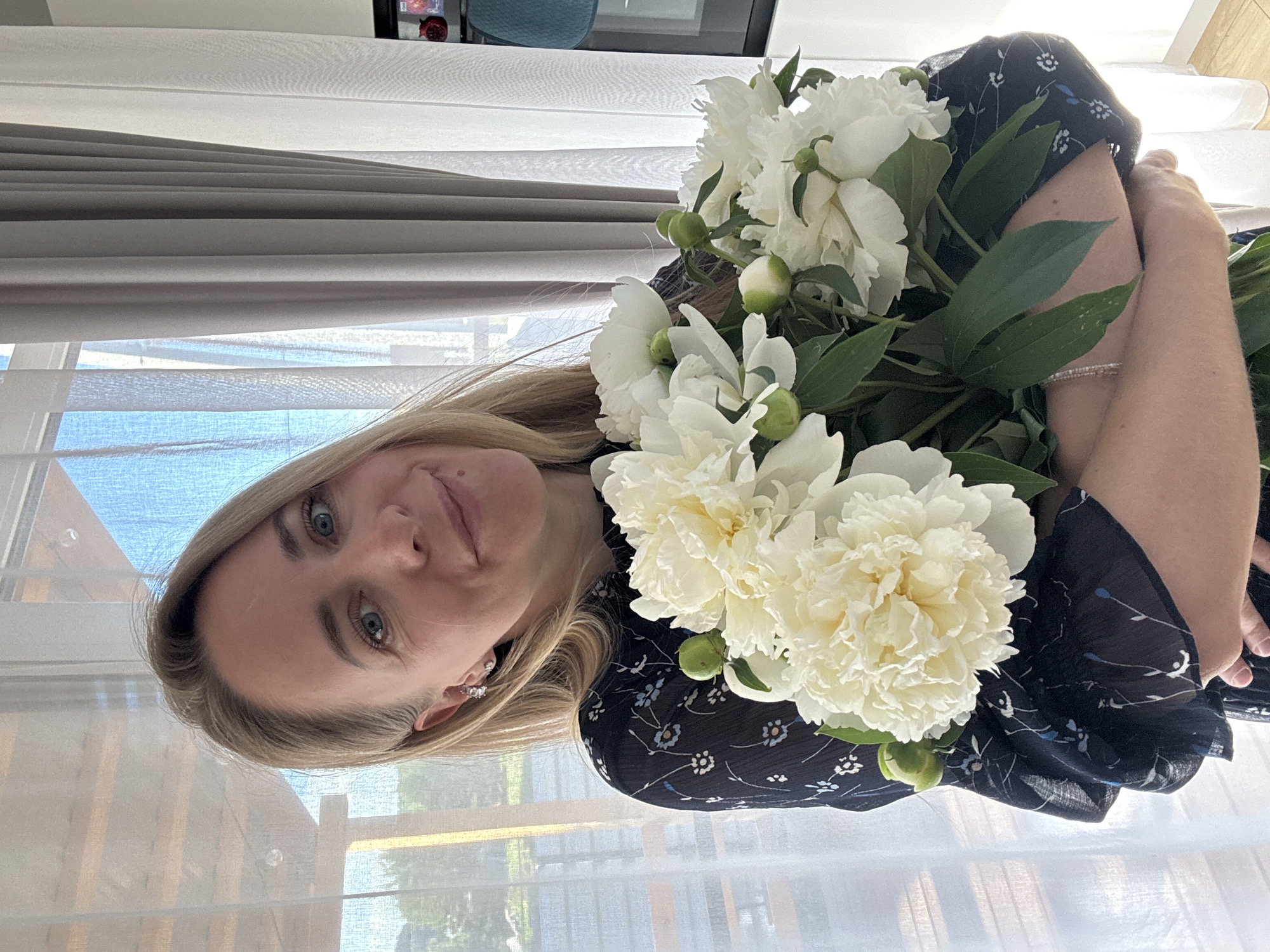 A woman with blonde hair and blue eyes, wearing a dark floral dress and silver earrings, is holding a bouquet of white peonies. She is smiling and looking at the camera. The background shows a window with sheer white curtains and a glimpse of the sky. The setting appears to be indoors, possibly a home in Chaikovsky, Russia, given the natural light and domestic ambiance.