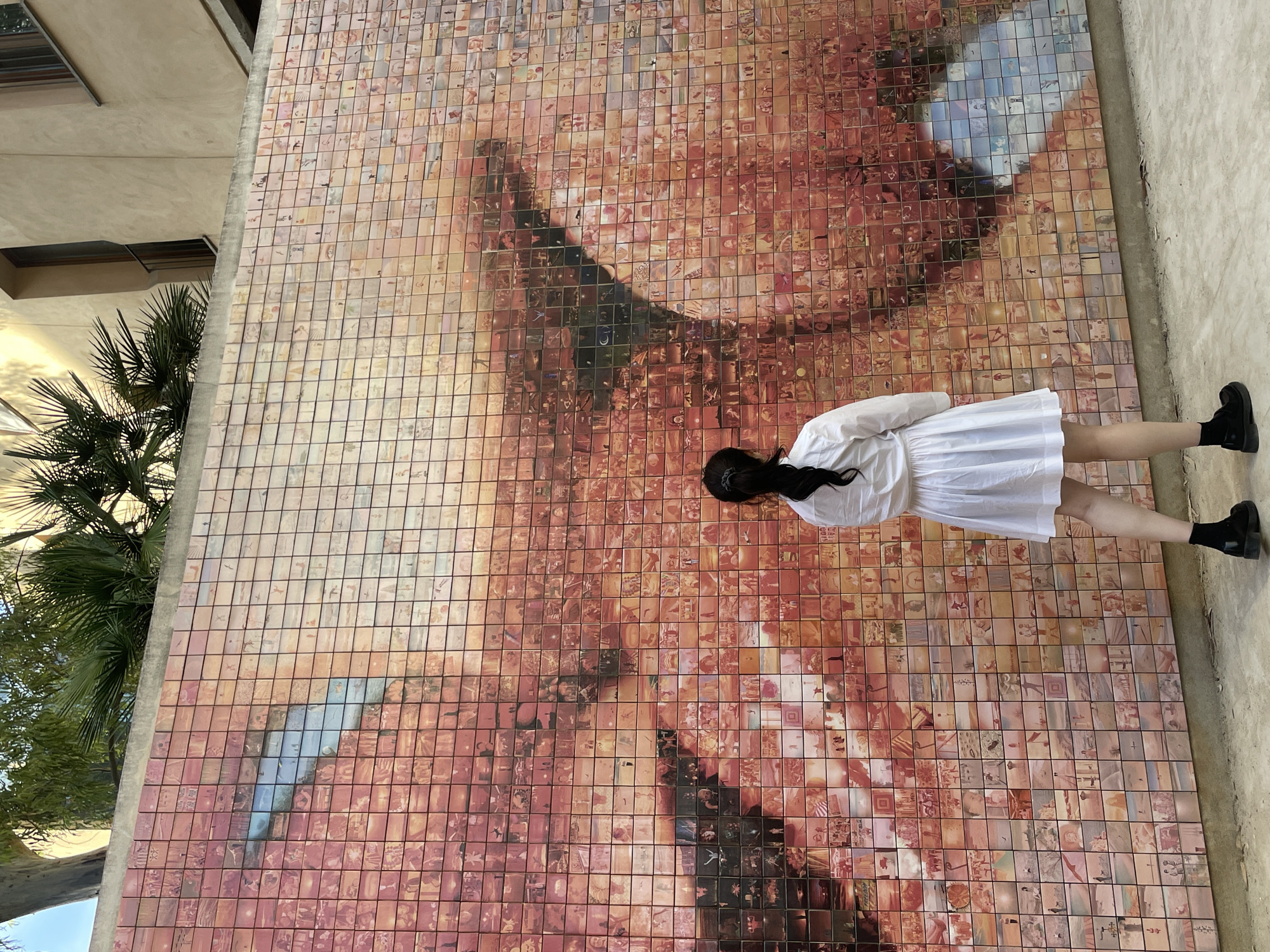 A person, observed from behind, stands facing a large outdoor mosaic wall in Plaça d'Isidre Nonell, Barcelona, Spain. The individual wears a white long-sleeved dress, black ankle boots, and black socks. Their dark hair is pulled back into a ponytail. The mosaic wall is constructed from numerous small, individual ceramic tiles, which collectively form a large-scale image of a human eye and a portion of a face. The dominant coloration of the mosaic ranges from red, orange, and brown tones, with a prominent blue section representing the iris and a tear-like streak. Each tile within the mosaic contains a distinct smaller image, contributing to the overall larger composite picture. Above the mosaic, a concrete ledge is visible, behind which the foliage of a tall eucalyptus tree and several palm trees protrude. To the upper right, a light-colored building with recessed windows is partially visible. The ground in the foreground where the person stands is paved with a light-colored, textured surface, likely concrete. The scene is illuminated by natural daylight.