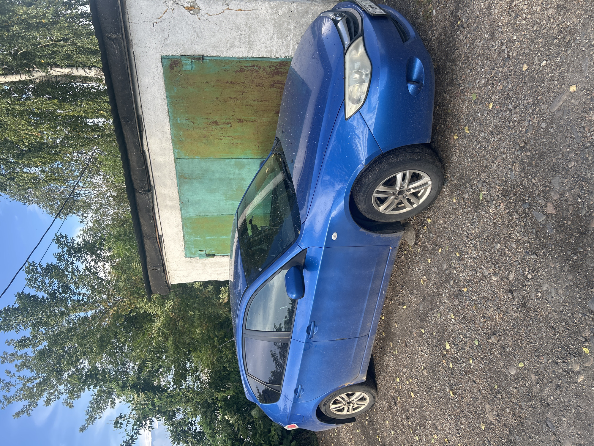 A blue Subaru Impreza hatchback is parked on a gravel surface in front of a rustic garage. The garage has a rough, white-plastered exterior with a large, weathered green metal door. Above the garage door, a dark, possibly tar-covered roofline is visible. The car is positioned facing towards the right side of the frame, with its front end slightly angled towards the garage. The background features lush green trees and a clear blue sky with some scattered clouds. A power line is visible running across the top of the image. The scene appears to be in an outdoor, possibly a rural or suburban, area. The car shows signs of dust and light debris, consistent with being parked outdoors. There are no people or other vehicles visible in the image.