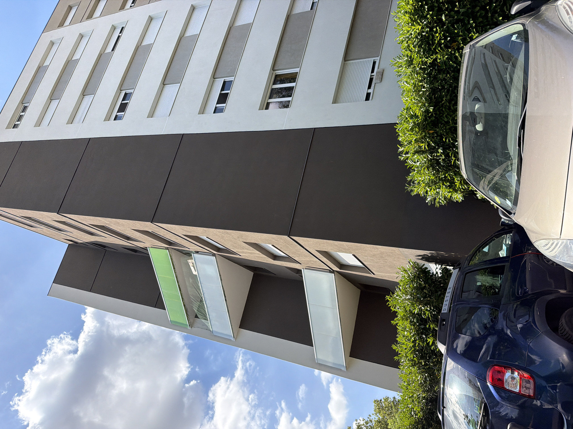 The image displays a modern apartment building with a dark grey facade and light beige sections featuring vertical white blinds and windows. The building has protruding balconies with frosted glass panels, one of which is a distinctive green color. In the foreground, partially visible cars are parked, one a silver sedan and another a dark blue SUV with "DUSTER" written on its rear. Lush green bushes flank the building and cars, suggesting an outdoor setting. The sky is a clear blue with scattered white clouds, indicating fair weather. The upward angle of the shot and the presence of cars and vegetation suggest an urban or suburban environment. The lighting implies it is daytime. No people are visible. The image is taken from a slightly tilted perspective.