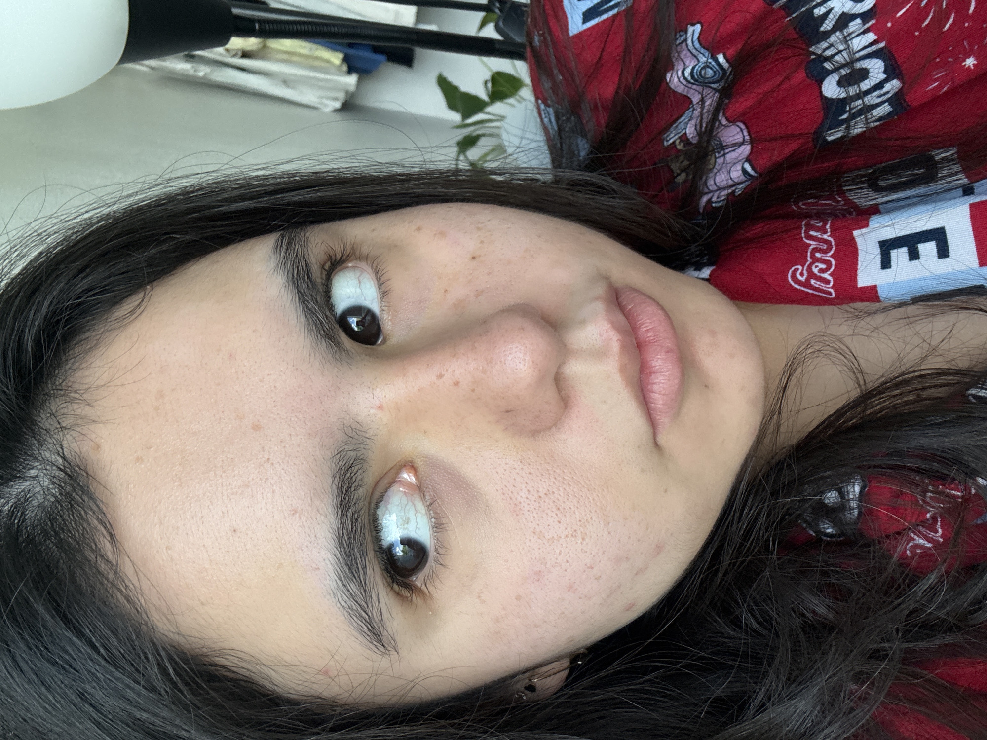 A close-up portrait displays a young person with long, dark hair, lying down and looking directly at the camera with a wide-eyed expression. The image is rotated, showing the face tilted to the left. The subject has dark irises, and the whites of their eyes are prominent, particularly the lower sclera. Their skin exhibits natural texture with visible pores and several small freckles or beauty marks across the forehead, nose, and cheeks. The lips are a natural pinkish hue, slightly parted.
The person is wearing a red garment, likely a casual top or pajamas, featuring white and blue patterns and text. Visible partial text on the clothing includes the letter "E", the letters "rry", and "ON", along with a stylized light pink/purple character.
The indoor background includes a white spherical object (likely a lamp or light fixture) and a black stand on the top left. Stacked papers or documents are also visible in this area. Towards the top right, a green leafy plant is partially seen. The setting suggests a home environment, possibly a bedroom or study, located in Austin, United States of America. The time of day is not explicitly indicated by the indoor lighting.