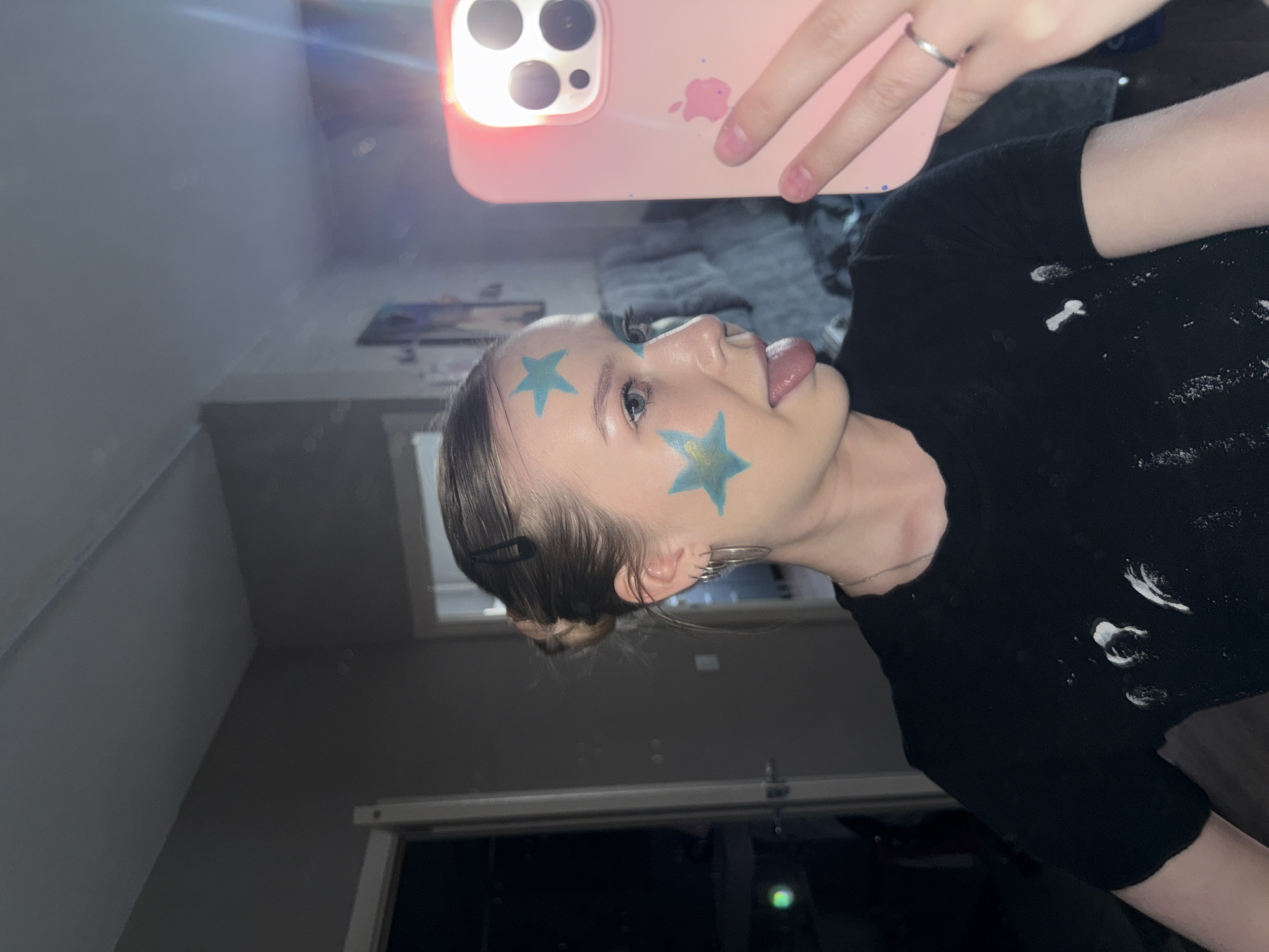 A person with stars painted on their face is taking a selfie. The person is wearing a black shirt. They are sticking out their tongue. They are indoors in a room that appears to be in a residential building. The photograph was taken near a window, which is visible. It is nighttime outside. I have no information about the photograph's location in the city of Chaikovsky, Russia.