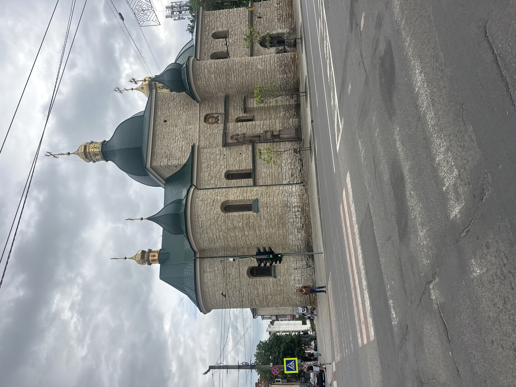 The image shows a large, stone church with green domes and golden crosses, located on a city street in Ternopil, Ukraine. The church has a classic architectural style with arched windows and decorative elements. In the foreground, there is a pedestrian crossing marked on the asphalt road. A traffic light is visible, showing a green signal. Several people are present in the scene, including individuals walking on the sidewalk and crossing the street. The sky is overcast with clouds. The overall environment suggests an urban setting with a mix of historical architecture and modern infrastructure.