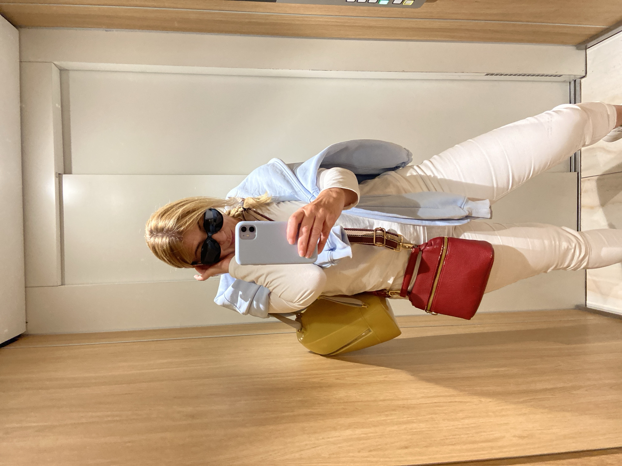 A woman is taking a selfie in an elevator. She is wearing sunglasses, a light blue hoodie, a white long-sleeved shirt, and white pants. She has two bags: a yellow bag and a red bag. The elevator has wooden floors and white walls. The control panel of the elevator is visible at the top of the frame. It is not possible to determine if the location is in the city of Budva, Montenegro based on this picture alone.
