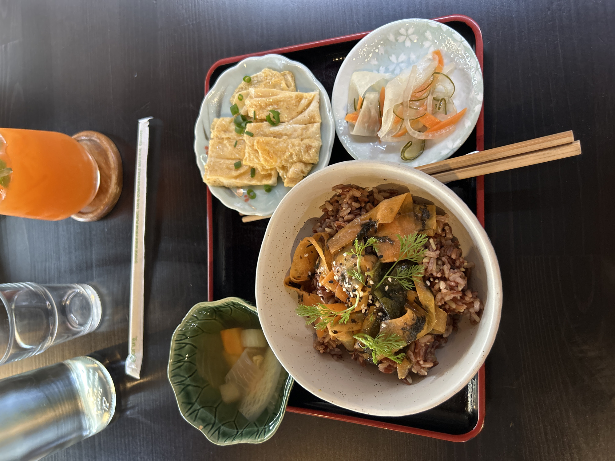 The image shows a meal served on a dark wooden table. The meal is presented on a red and black tray. On the tray, there is a white bowl filled with brown rice topped with cooked vegetables, possibly carrots and greens, and sprinkled with sesame seeds and dill. To the right of the bowl are two small plates. One plate contains folded pieces of what appears to be cooked egg or tofu, garnished with chopped green onions. The other plate holds a selection of pickled vegetables, including sliced cucumbers, carrots, and radishes. A small green bowl with a decorative pattern contains a clear broth with pieces of vegetables. In the background, there is a glass bottle of water and an empty drinking glass. Next to them, a glass of orange juice sits on a wooden coaster. A white paper straw is placed on the table. The overall scene is of a prepared meal, likely in a restaurant setting in Chiang Mai City Municipality, Thailand. There are no people visible in the image.