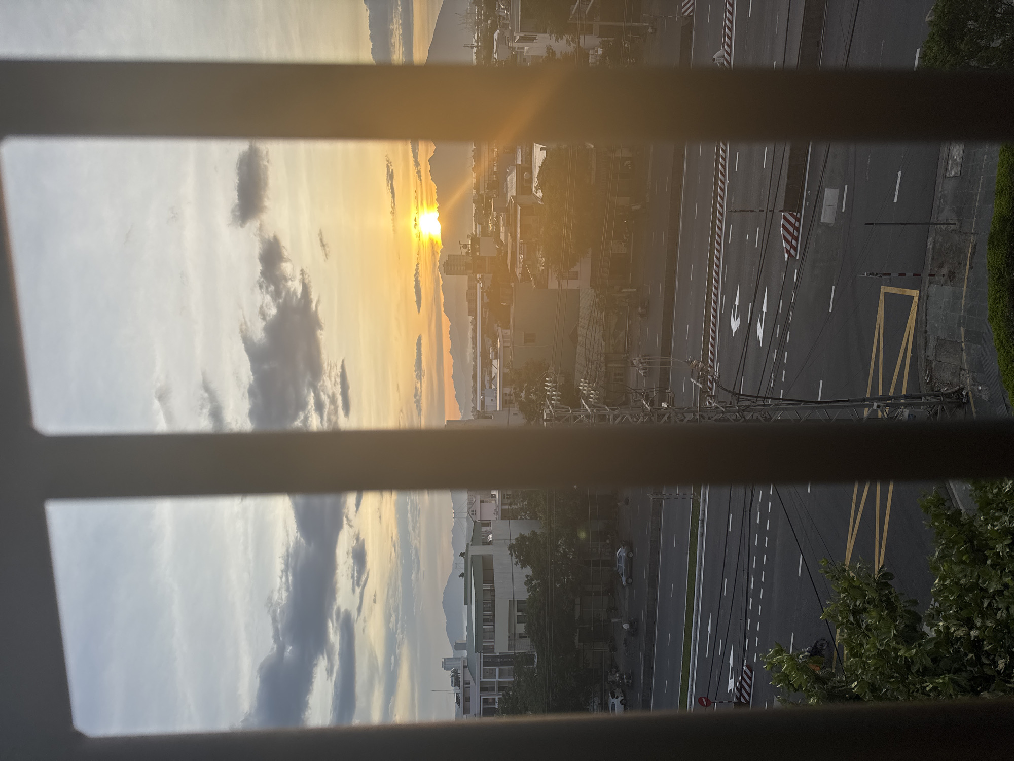 This is a photograph taken from behind a set of vertical bars, looking out over a city street at sunset. The view is framed by the bars, which are a dark color. In the distance, the sun is setting behind a range of mountains, casting a warm orange and yellow glow across the sky and the cityscape. Clouds are scattered across the sky, some appearing dark and dramatic, while others are lighter and wispy.
The city below appears to be a mix of low-rise buildings and taller structures. Trees line some of the streets, and there are visible power lines and utility poles. A multi-lane road runs horizontally across the middle of the frame, with cars and motorcycles visible on it, as well as road markings including white and yellow lines and directional arrows. There are also traffic signs and barriers visible along the road. The scene suggests a fairly urban environment. Given the mountain range in the background and the coastal city setting, this is likely Đà Nẵng, Vietnam.
