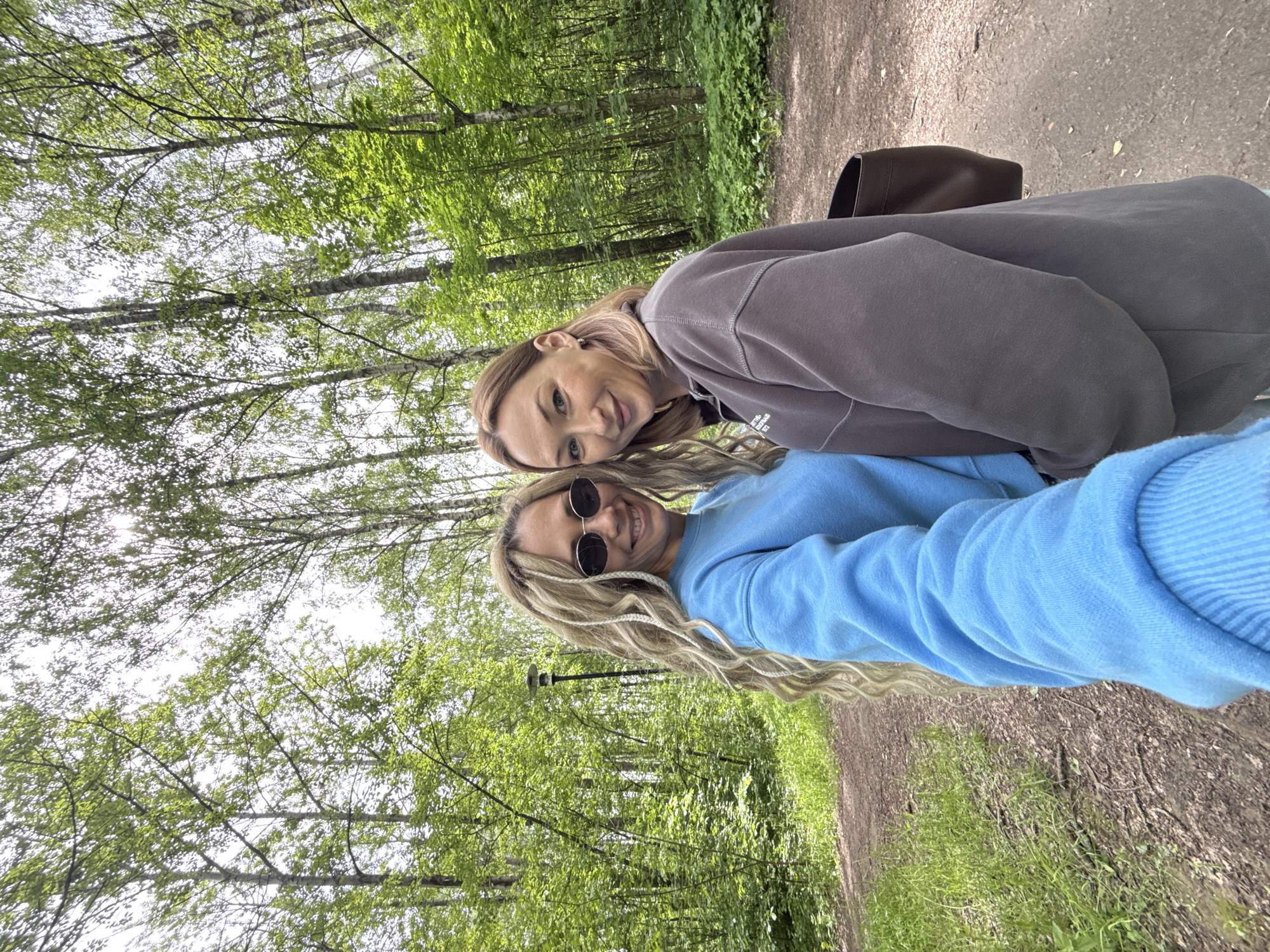 Two women are posing for a selfie on a path surrounded by trees. One woman wears a blue hoodie and sunglasses, while the other wears a gray hoodie. A brown bag is visible behind the woman in gray. The location is Rusanovo, Russia.