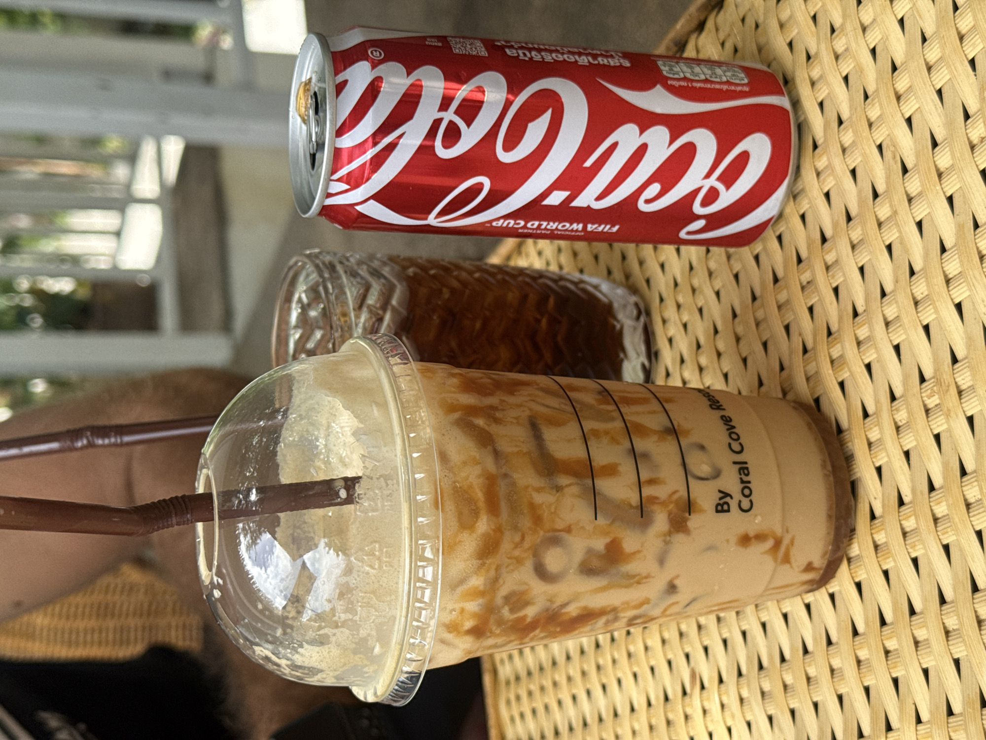 The image captures a close-up perspective of beverages on a woven bamboo tray. In the foreground, a clear plastic cup contains an iced coffee with caramel swirls and a brown plastic straw. The cup has markings indicating it is "By Coral Cove Res". To the right of the coffee cup is a red Coca-Cola can, open, with its silver pull-tab visible. Behind the coffee cup and to its left, a patterned glass filled with a dark liquid, presumably iced tea or coffee, is partially visible. The background shows hints of outdoor seating with a person's torso and arm, suggesting an open-air cafe or restaurant environment. The lighting indicates it is daytime. The provided context suggests this scene is located in the Ko Samui District of Thailand, likely at a restaurant associated with Coral Cove.