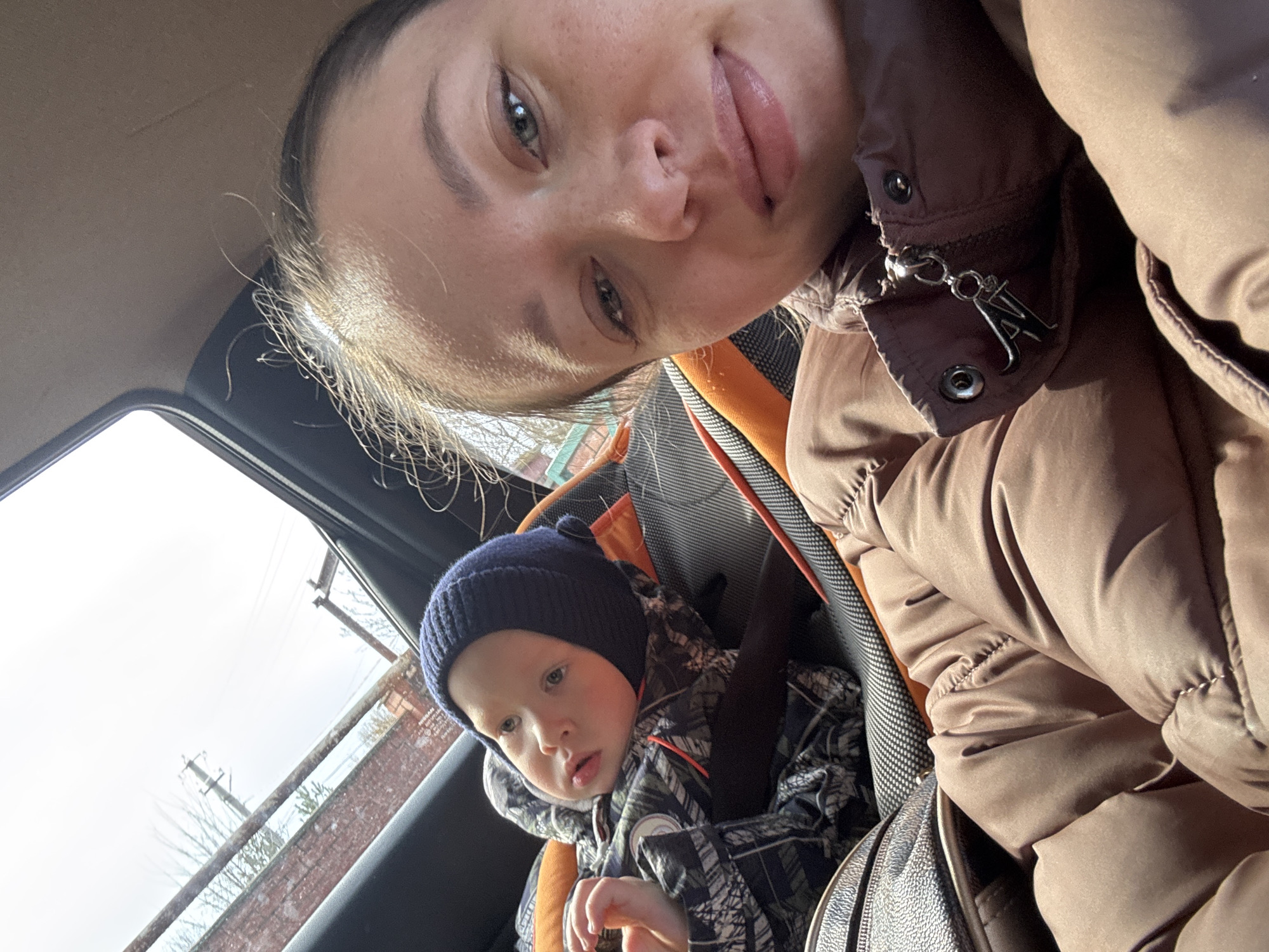 An adult female and a young child are observed inside a vehicle in the city of Ферма, Russia. The female, positioned on the right, faces the camera for a selfie, wearing a dark brown puffer jacket with a zipper featuring an "A" design. She has light skin, visible freckles, and dark hair pulled back. The child, seated in a black and orange car seat to the left of the female, wears a dark blue knitted beanie and a camouflage-patterned winter jacket. The child's gaze is directed slightly upwards and to the left. A dark, textured bag is partially visible in the lower-left foreground. Through the vehicle's sunroof, an exterior scene includes a reddish-brown brick wall, bare tree branches, utility poles, and overhead power lines against a bright, possibly overcast sky.