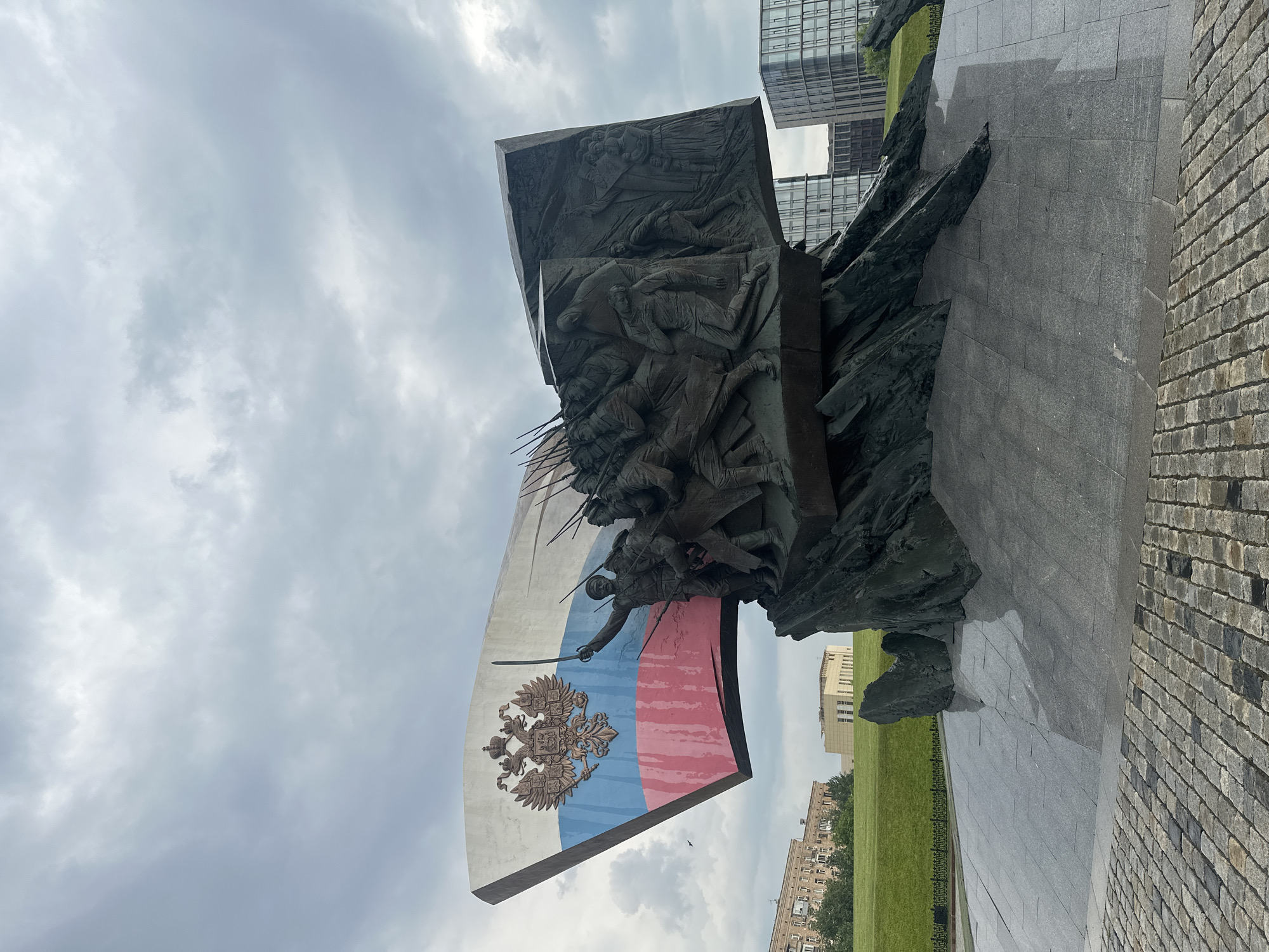 The image shows the "Monument to the Defenders of Moscow" located on Poklonnaya Hill in Moscow, Russia. The monument features a large, stylized Russian flag made of metal, with the double-headed eagle emblem prominently displayed. Below the flag, a dramatic bronze relief depicts soldiers in battle, charging forward with bayonets. The relief is set against a jagged, rock-like base. In the background, modern glass-fronted buildings rise against a cloudy sky. The foreground shows a paved area with cobblestones, and a manicured green lawn stretches out behind the monument. The overall impression is one of solemn remembrance and patriotic pride, set within an urban landscape.