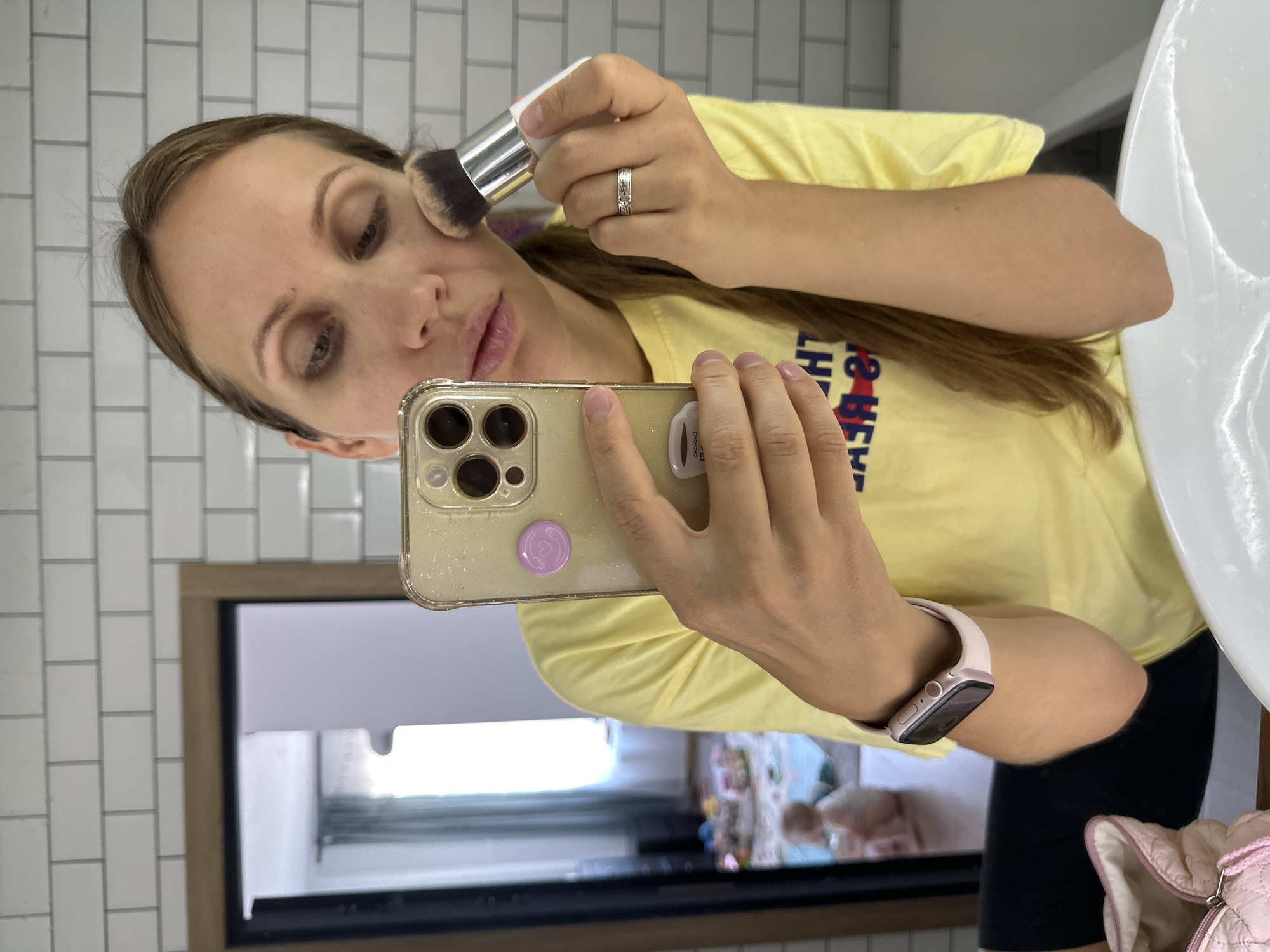 A woman with long brown hair is applying makeup using a large brush to her face, holding a smartphone in her left hand. She is wearing a yellow t-shirt with text and a pink smartwatch on her left wrist. She is standing in a bathroom with white subway tiles on the wall and a mirror reflecting a room with a child playing.