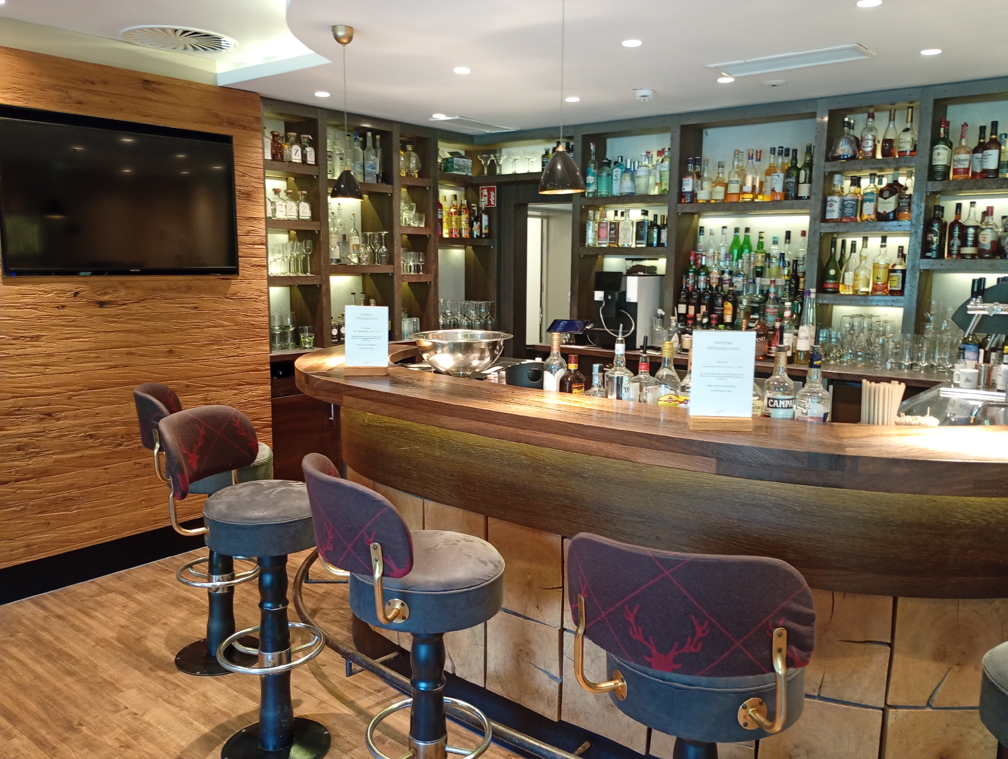 The image depicts the interior of a bar, likely within a hotel in Bad Sachsa, Germany, given the context provided. The bar area is modern and features a prominent wooden counter with a curved design. Behind the counter, shelves are stocked with a wide variety of liquor bottles, glassware, and other bar supplies, illuminated by warm lighting. Several bar stools with upholstered seats and backs, featuring a distinctive red stag pattern, are arranged around the counter. A large flat-screen television is mounted on a wooden-paneled wall to the left of the bar. The flooring appears to be wooden. The overall ambiance suggests a sophisticated and well-equipped establishment.