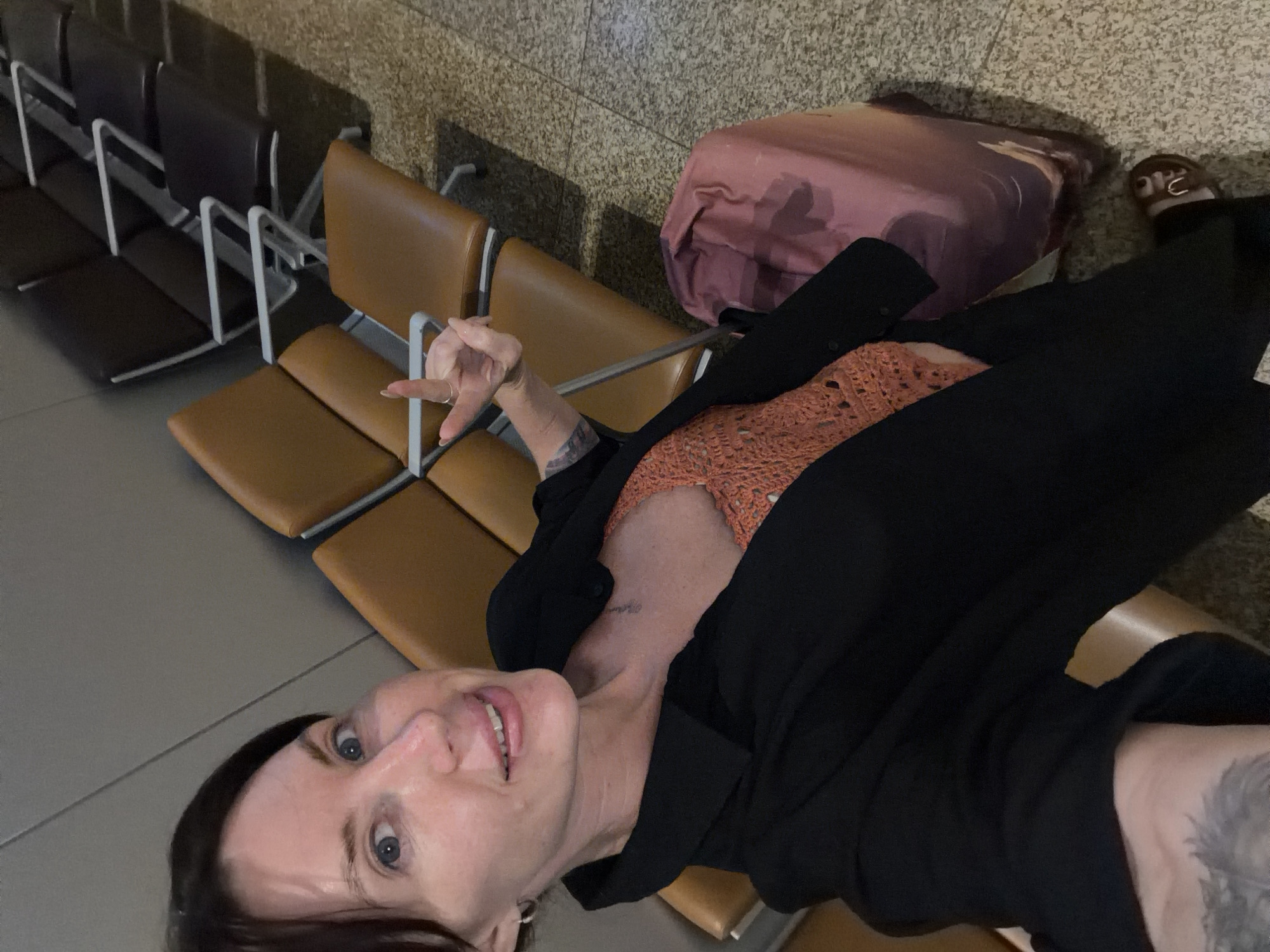 A woman is taking a selfie in what appears to be an airport waiting area, possibly in Bac Cam Ranh Ward, Vietnam. She has dark hair pulled back and is wearing a black jacket over an orange crocheted top. She is smiling and holding up her left hand, showing off a ring. To her right, a pink suitcase is visible. The background consists of rows of empty brown airport chairs and speckled flooring.