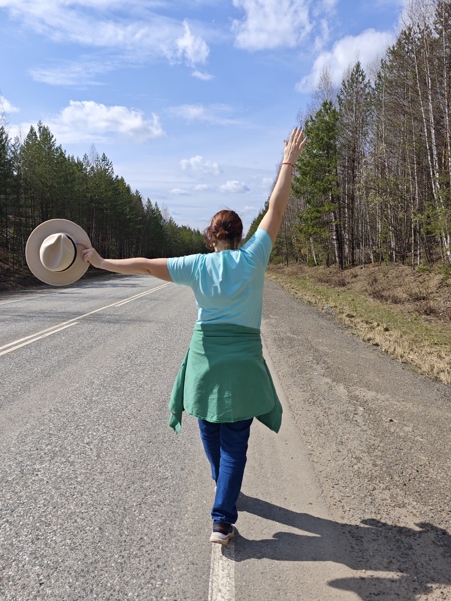 A woman stands on a road in Chaikovsky, Russia, with forests bordering both sides. She faces away from the viewer, wearing jeans, a light blue t-shirt, and a green shirt tied around her waist. Her dark hair is pulled back. Her left arm is extended, holding a beige hat. Her right arm is raised in the air. The road is grey with white lines, and the sky above is blue with scattered white clouds.