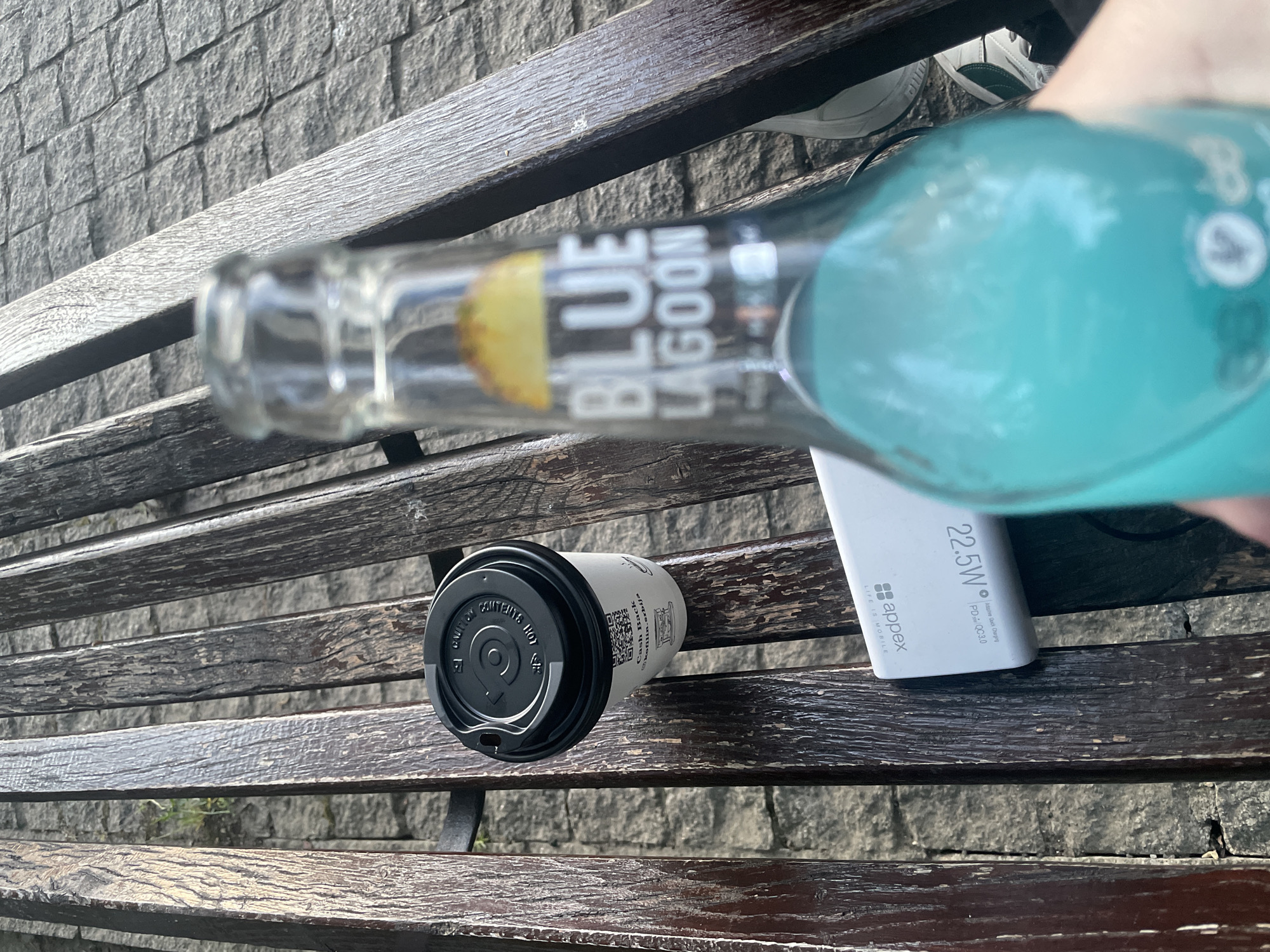 The image presents an overhead, angled view of various items resting on a weathered wooden slatted bench, suggesting an outdoor public setting in Novi Sad, Serbia. A hand is holding a glass bottle containing a bright turquoise liquid, identified by its label as "BLUE LAGOON" with a yellow fruit graphic.
Adjacent to the bottle, a white power bank is visible, bearing the brand "appex" and the slogan "LIFE IS MOBILE." Technical specifications such as "22.5W," "Adaptive Quick Charging," and "PD+QC3.0" are clearly printed on its surface.
Further down the bench, a white paper coffee cup with a black lid is placed. The cup features a QR code, the text "Cash Back," and "kofilin.srbija." The lid also displays partially visible warnings like "CAUTION" and "CONTENTS HOT."
In the upper right corner, a portion of a foot wearing a white and dark green sneaker is visible. The background consists of a greyish-brown stone wall. The bright, natural lighting indicates it is daytime. The overall scene depicts a casual moment of rest, possibly with a refreshing drink, coffee, and a device being charged.