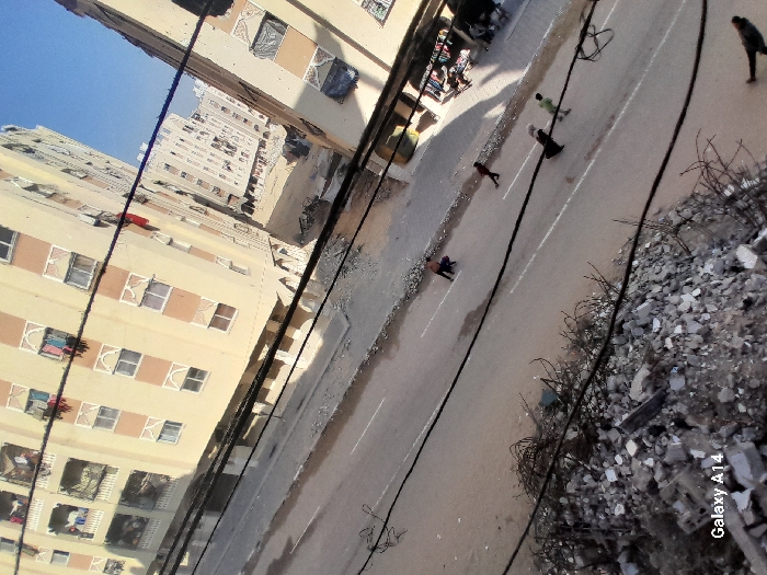 An elevated, tilted view captures a street scene in the city of Khan Yunis, Palestinian Territory. In the immediate foreground, a substantial pile of concrete rubble and exposed rebar occupies the bottom left. Multiple black utility cables crisscross the upper portion of the foreground. A paved road with a white broken center line runs diagonally through the middle ground. Debris is scattered along the road's edges.
Seven individuals are present on the road. One person, dressed in dark clothing, is crouched in the left lane. To their right, another individual in dark clothing walks. Further right, a group of three individuals walks together: one adult in a dark, long garment and two smaller individuals, one in a light green top and the other in dark attire. A seventh individual is partially visible walking towards the right in the bottom right corner.
Multi-story residential buildings line both sides of the street. The prominent building on the left is light-colored with beige and white facades, featuring multiple windows with decorative frames and balconies from which laundry hangs. A market stall or shop with items displayed is visible beneath the building on the right side of the street. In the background, further urban structures are visible under a clear blue sky. The scene is illuminated by daylight, with visible shadows. A "Galaxy A14" watermark appears in the bottom left.