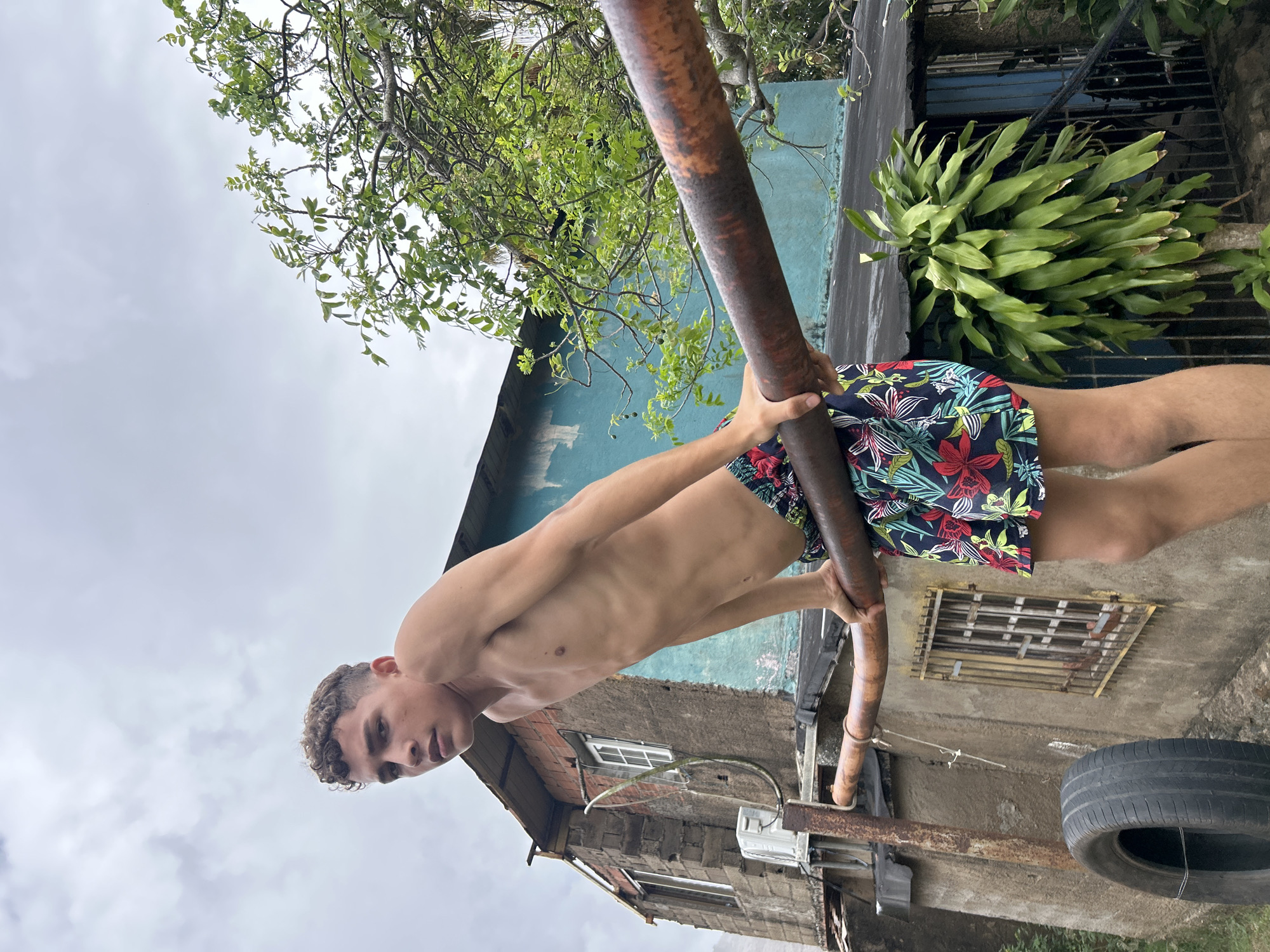 A young man with short, curly brown hair and a toned, shirtless physique is performing a calisthenics move, holding onto a rusty metal bar. He is wearing colorful swim shorts with a floral pattern featuring red, white, blue, and green motifs. His gaze is directed towards the viewer, with a serious expression.
The setting appears to be outdoors in what looks like a residential area, possibly a backyard or an alley. The background includes parts of old, weathered buildings made of concrete or brick, some with visible windows and metal grates. One building wall is painted a light blue. There are lush green plants and tree branches with leaves visible, suggesting a natural environment alongside the urban elements.
The sky above is overcast with a light gray tone, indicating a cloudy day. The time of day is not explicitly clear but the lighting suggests daytime. There's an old tire lying on the ground near the lower right of the frame, and some additional rusty metal structures are visible. The image is oriented vertically, and the man is positioned diagonally across the frame, giving the impression of an energetic movement or pose.
No visible text is present in the image. The overall atmosphere is one of strength and focus amidst a somewhat rugged and natural backdrop, typical of outdoor fitness routines in diverse locations. Given the location context of Pedro González, Venezuela, this scene aligns with the typical architectural styles and natural vegetation that can be found in the region.