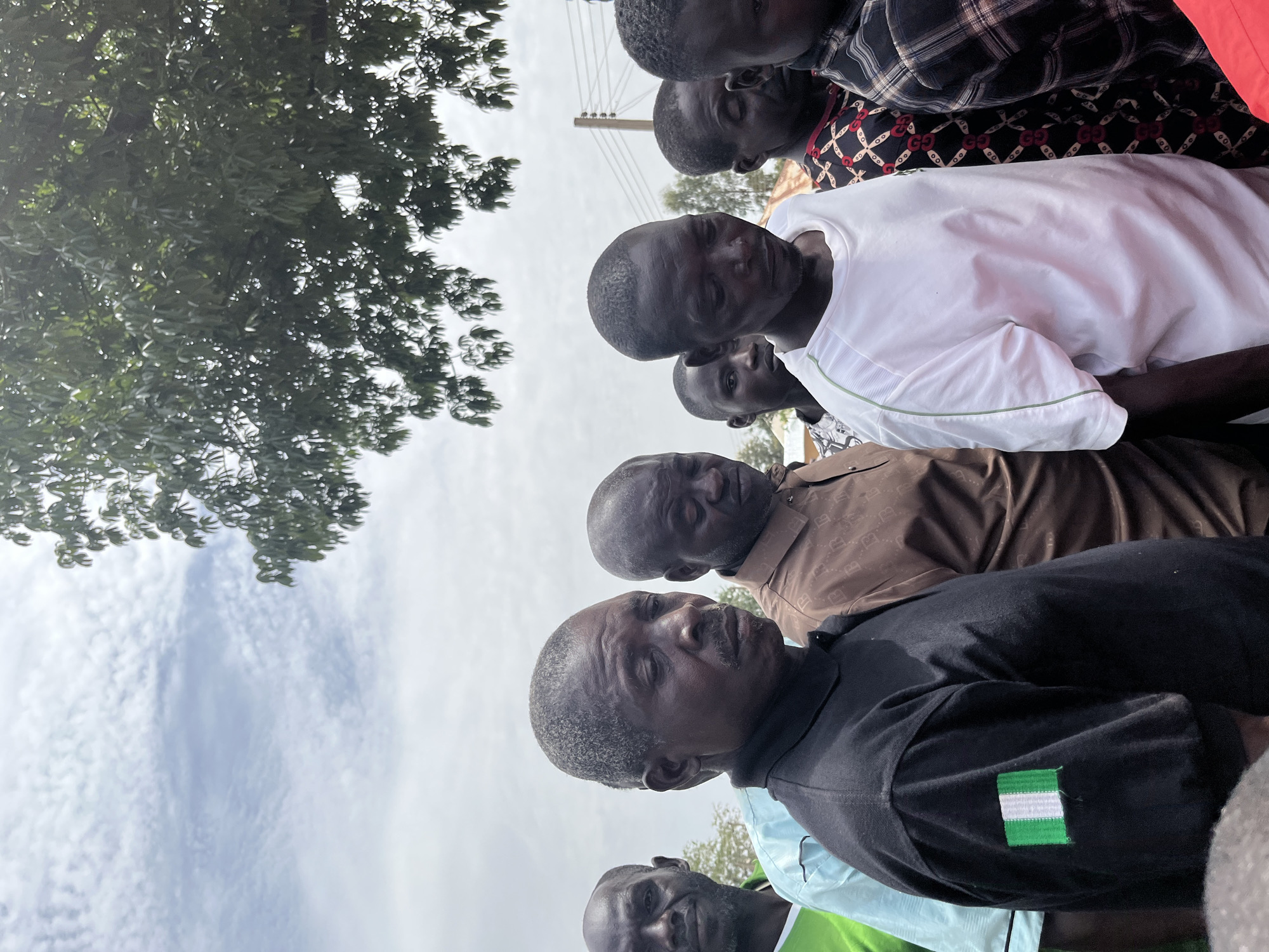 A group of seven or more Black men are seen in Dutsen Kato, Nigeria. They are standing closely together, facing forward. The men are wearing casual clothing, including a white t-shirt, a brown button-down shirt, and a black polo shirt with a Nigerian flag patch. A green tree and cloudy sky are visible in the background.