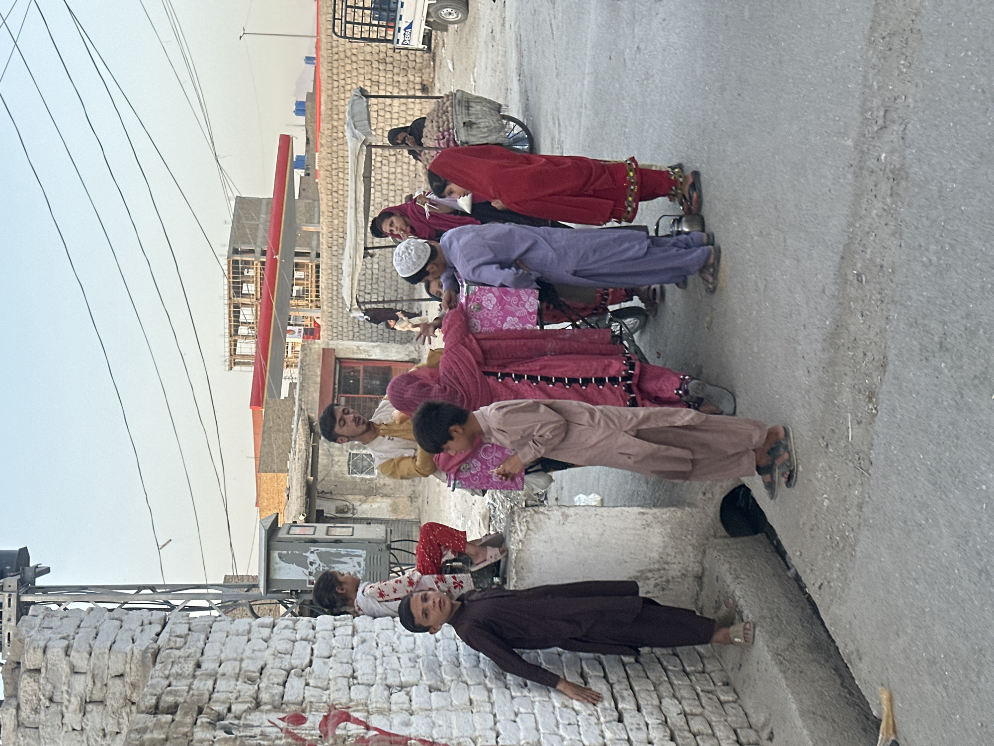 In Quetta City Tehsil, Pakistan, a street scene unfolds with a group of people, predominantly children and some adults, gathered near a brick wall. The environment suggests a residential or market area with a mix of structures, including a partially built building in the background, a power pole with overhead wires, and a cart laden with produce, possibly potatoes. The people are dressed in traditional South Asian attire. A young boy in a brown shalwar kameez stands by the wall, while other children and adults are engaged in various interactions. Some appear to be looking at a pink box or bag, possibly receiving or exchanging items. A woman in a red headscarf is visible near the cart, and another woman in a pink headscarf is also present. The ground is paved with asphalt and concrete, with a visible drainage ditch running along the side. The lighting indicates it is likely daytime, with a clear sky.