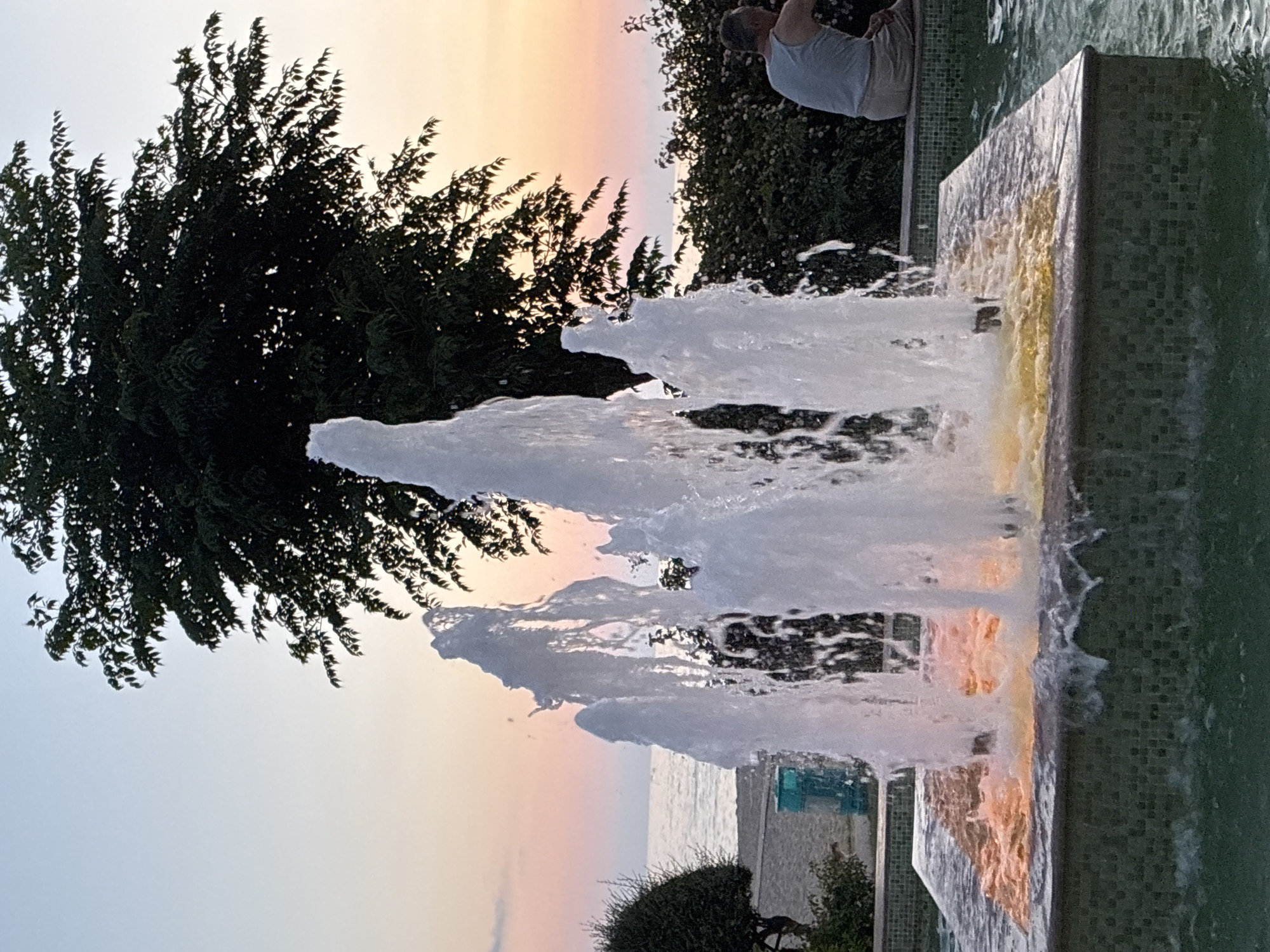 A fountain with multiple jets of water is situated in the foreground. The fountain's basin is tiled with small green mosaic tiles, and the water within it is illuminated with an orange glow. In the background, a large tree with dark green leaves is silhouetted against a twilight sky. The sky displays soft gradients of blue and pink. To the right of the fountain, a person wearing a light blue tank top and light-colored shorts is seated, facing away from the camera. The background also shows a body of water, likely the sea, with a beach and some vegetation. The scene appears to be located in Alanya, Turkey, possibly near the coastline.