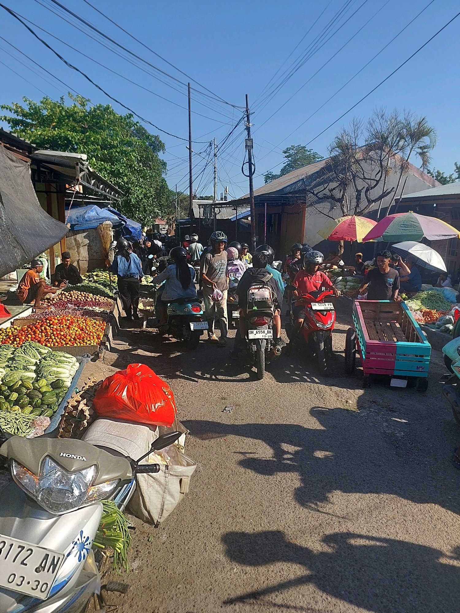 This is a street scene in Kupang, Indonesia, likely a market area. The image captures a sunny day with clear blue skies. In the foreground, a silver Honda scooter is parked, its license plate visible as "3172 AN 03 30". Behind the scooter, a large red plastic bag is placed on a light-colored canvas bag, which is itself on the scooter's seat. To the left of the scooter, a display of fresh produce is laid out, including green beans and what appear to be cucumbers. Further back, more vegetables, including what look like tomatoes and onions, are piled high in various containers.
Several people are present, most of whom are on or near motorcycles. A person wearing a blue denim jacket is walking away from the viewer, holding a broom. Another person in a red shirt is riding a red motorcycle. A man in a black t-shirt is standing next to a colorful wooden cart with blue and pink accents, which is attached to a motorcycle. Other individuals are visible in the background, some on motorcycles and some standing near stalls. The street is lined with temporary structures, some covered with tarps, suggesting market stalls. Power lines crisscross above, supported by utility poles. The overall atmosphere is that of a busy marketplace with ongoing transactions and movement.