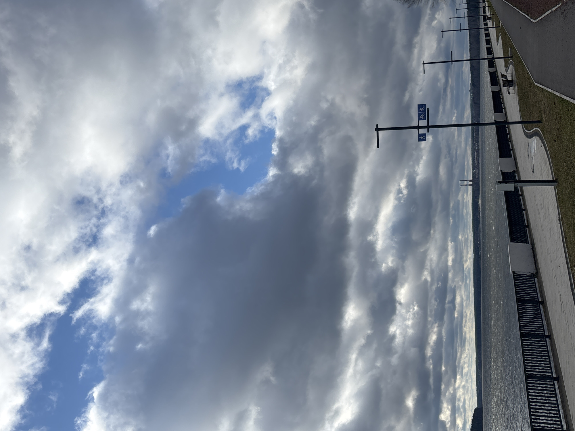 The image captures a scenic waterfront promenade in Chaikovsky, Russia, under a dramatic, partly cloudy sky. The photograph is taken from a slightly elevated perspective looking along the water's edge.
The sky occupies a significant portion of the frame, filled with a dynamic display of grey and white clouds. Patches of bright blue sky are visible, particularly towards the left and center, indicating breaks in the cloud cover. The clouds closer to the horizon appear brighter, suggesting that sunlight is either behind them or breaking through.
A large body of water, likely a lake or wide river, stretches across the middle of the scene, its surface reflecting the light from the sky and appearing slightly rippled. Across the water, a faint, hazy landmass or distant shoreline is visible.
Along the right side of the image, a well-maintained promenade runs parallel to the water. It consists of a dark, paved path (likely asphalt) and an adjacent lighter-colored, textured path, possibly for walking or cycling, separated by a thin darker line. Next to the path is a strip of green grass. Lining the promenade are several tall, dark poles, each with horizontal crossbars. One of these poles, positioned prominently in the mid-ground, features two blue rectangular signs with white pictograms. The upper sign depicts a bicycle, indicating a bicycle path, while the lower sign shows a person walking, indicating a pedestrian path. Further along the promenade, dark benches are visible, suggesting places for rest.
The time of day appears to be daytime, with variable weather characterized by both bright light and significant cloud cover. No specific human activity is depicted; the scene is static, showcasing the infrastructure of the waterfront and the natural elements.