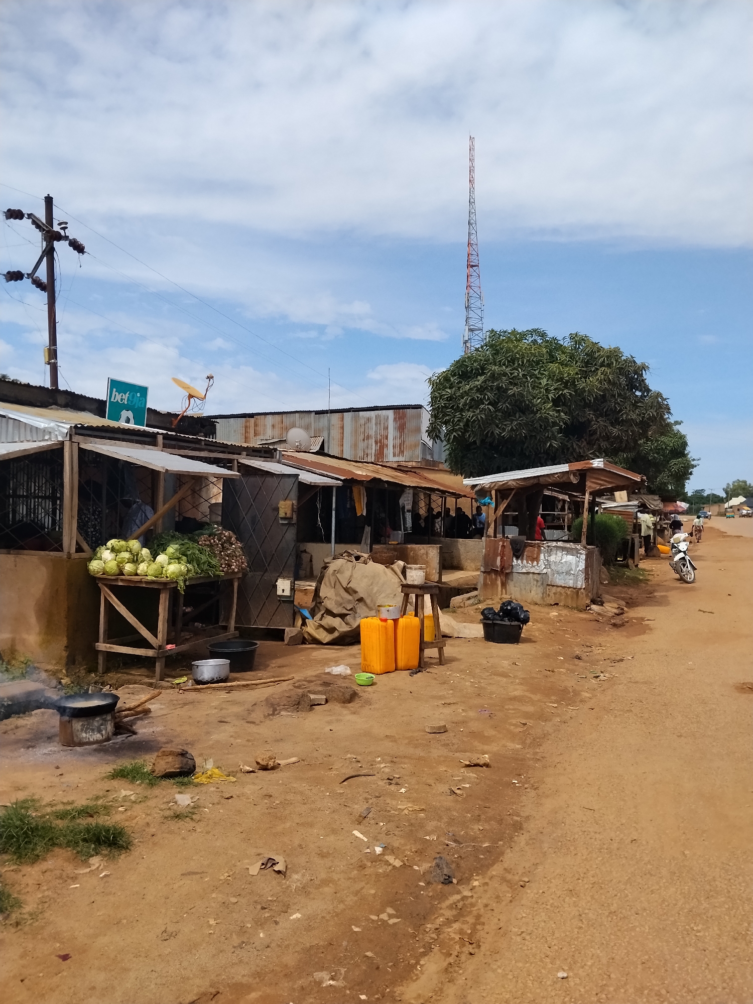 This image depicts a bustling market scene on a dirt road in Nabatum, Nigeria. The foreground features an outdoor cooking station with a pot on a fire, surrounded by scattered debris. To the left, a stall displays a variety of fresh produce, including cabbage and what appears to be onions, atop a wooden table. Several black and silver buckets, along with yellow water containers, are visible near the stall.
Further into the scene, a row of small shops with corrugated metal roofs line the road. These structures appear to be made of wood and metal, some with open fronts and others with gated entrances. In front of one shop, sacks are piled up. A motorcycle is parked to the right of the shops. People are visible inside and around some of the shops, suggesting ongoing commercial activity.
In the background, a large, leafy tree stands prominently, with a tall, lattice radio tower rising behind it against a cloudy blue sky. The road itself is unpaved, indicating a rural or semi-urban setting. The overall impression is one of vibrant, everyday commerce in a Nigerian community.