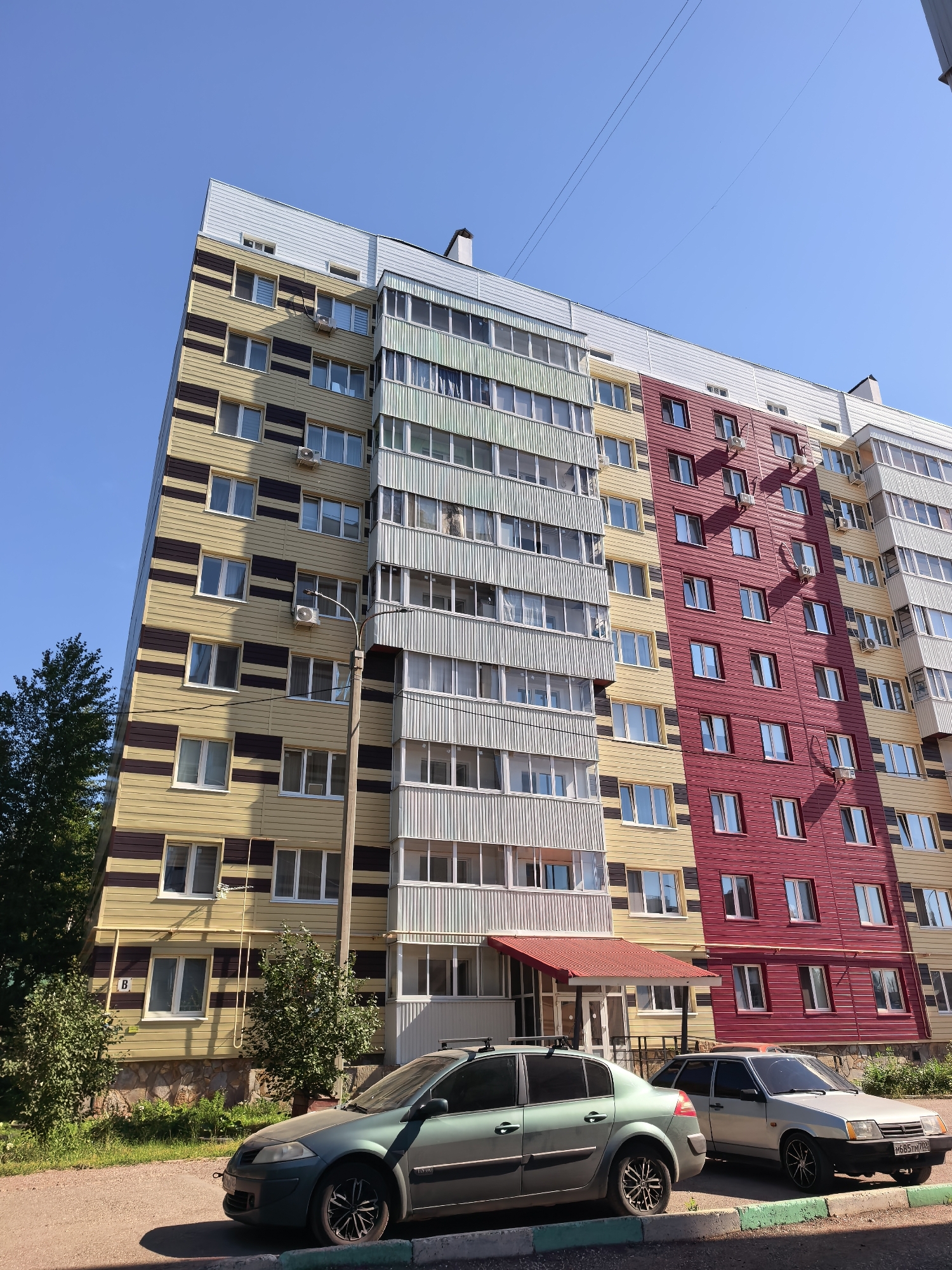 The image depicts a residential building with a distinctive facade featuring alternating horizontal bands of yellow and dark brown on the left section, a central section with vertical pale green and white stripes, and a right section with vertical red panels. The building has multiple stories with numerous windows and balconies. In the foreground, two cars are parked on a paved surface next to a curb. The car on the left is a greenish-blue sedan, and the car on the right is a silver hatchback. A small tree is visible to the left of the building, and a larger tree is on the far left. The sky is clear and blue. The building appears to be located in a residential area in Sterlitamak, Russia, as suggested by the car license plates, one of which reads "M685TN702 RUS". The scene is illuminated by sunlight, casting shadows.