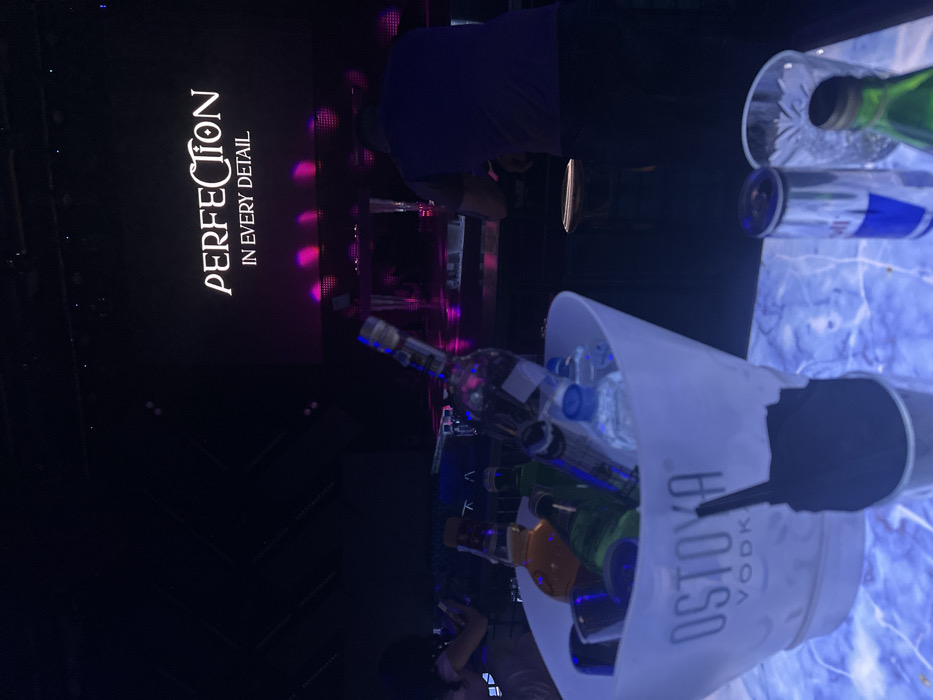 The scene depicts the interior of Perfection Club, a nightclub located in Varna, Bulgaria. The vantage point is from a patron's table or bar area, looking towards the stage.
In the immediate foreground, a white plastic ice bucket, branded "OSTOYA VODKA," rests on a table surface that emits a blue-white light and exhibits a marbled pattern. The bucket contains a large clear glass bottle of Ostoya Vodka, several smaller clear plastic water bottles, multiple green glass bottles, and one amber-colored bottle. A black napkin is positioned upright in a clear glass in front of the ice bucket. To the right of the bucket, on the same illuminated surface, an open silver Red Bull can, an empty cut-glass tumbler, and the neck of a green glass bottle are visible.
In the mid-ground, a male figure, seen from the back, wearing a dark purple t-shirt and dark pants, is standing at a counter or bar. To the far left, partially visible, is a female figure with dark, curly hair, wearing a light-colored garment, holding a smartphone in her hands.
The background features a large, dark stage screen displaying the text "PERFECTION" in a stylized white font, with "IN EVERY DETAIL" written below it in a standard white font. The stage area below the screen is illuminated with magenta and purple stage lights, reflecting off the dark surface. The overall environment is dimly lit, consistent with a nightclub atmosphere, with prominent artificial lighting sources.