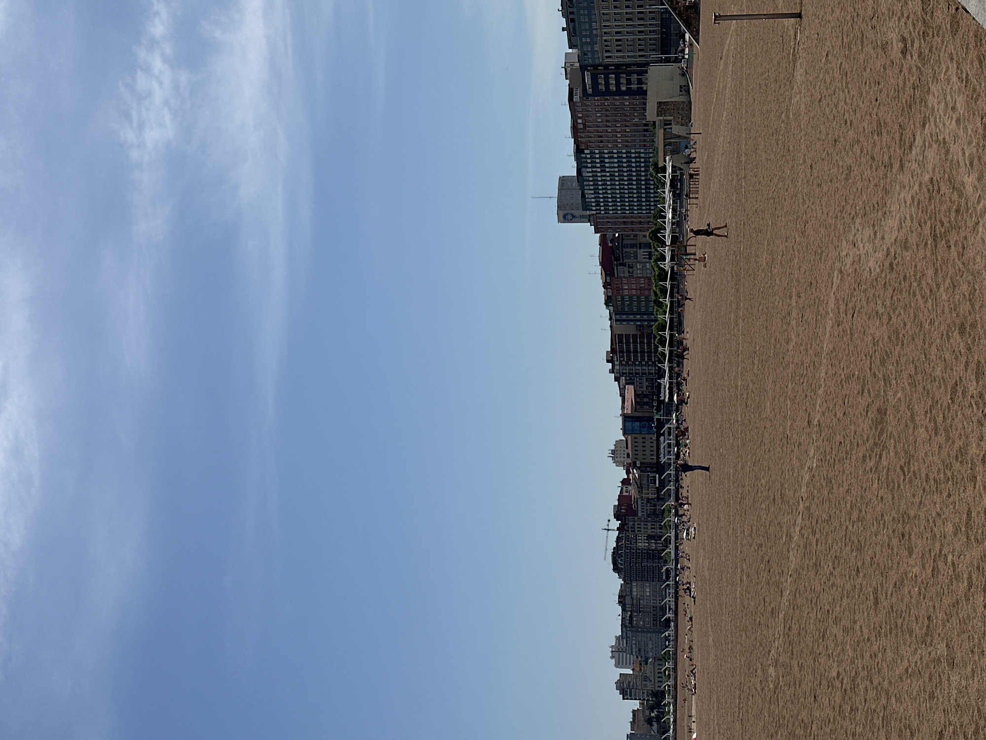 The image depicts San Lorenzo beach in Gijón, Spain, on a sunny day. The composition is horizontally oriented, with the beach stretching to the right and the city skyline on the left. The beach is crowded with people, and buildings line the coastline. The sky is mostly clear, with some clouds.