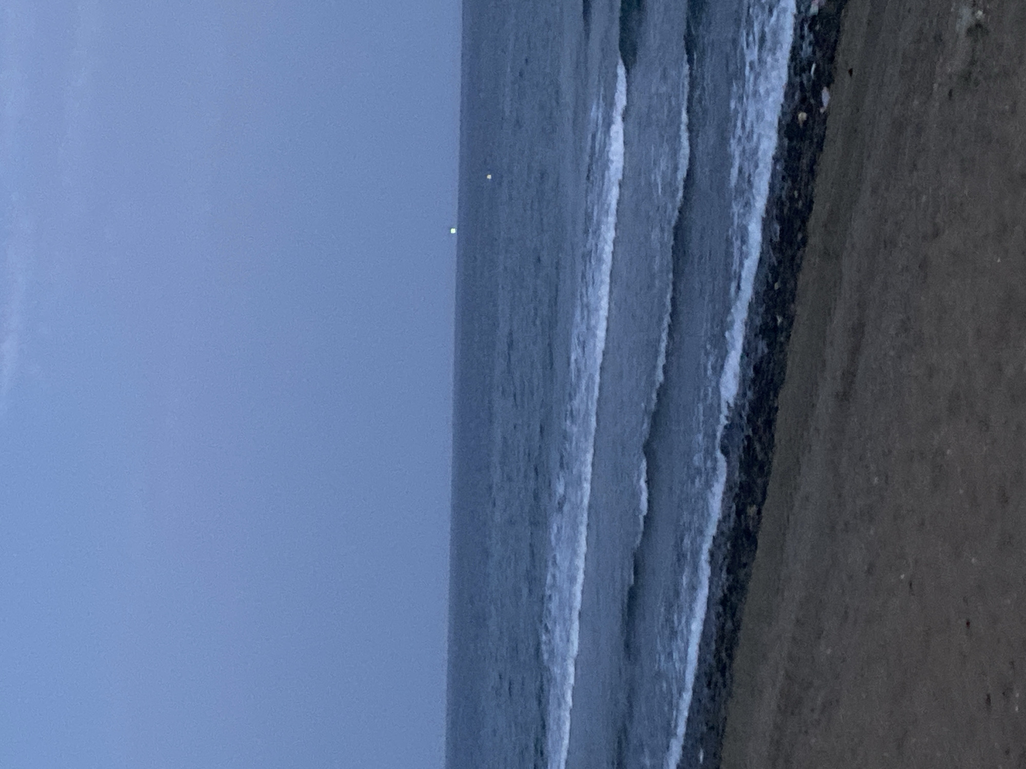 A view of the sea from a beach at dusk, with waves breaking on the shore. In the distance, there are two lights visible on the horizon. The sky is a deep blue, and the water is a dark grey. The beach in the foreground is made of sand and small pebbles. This scene depicts a tranquil coastal environment.