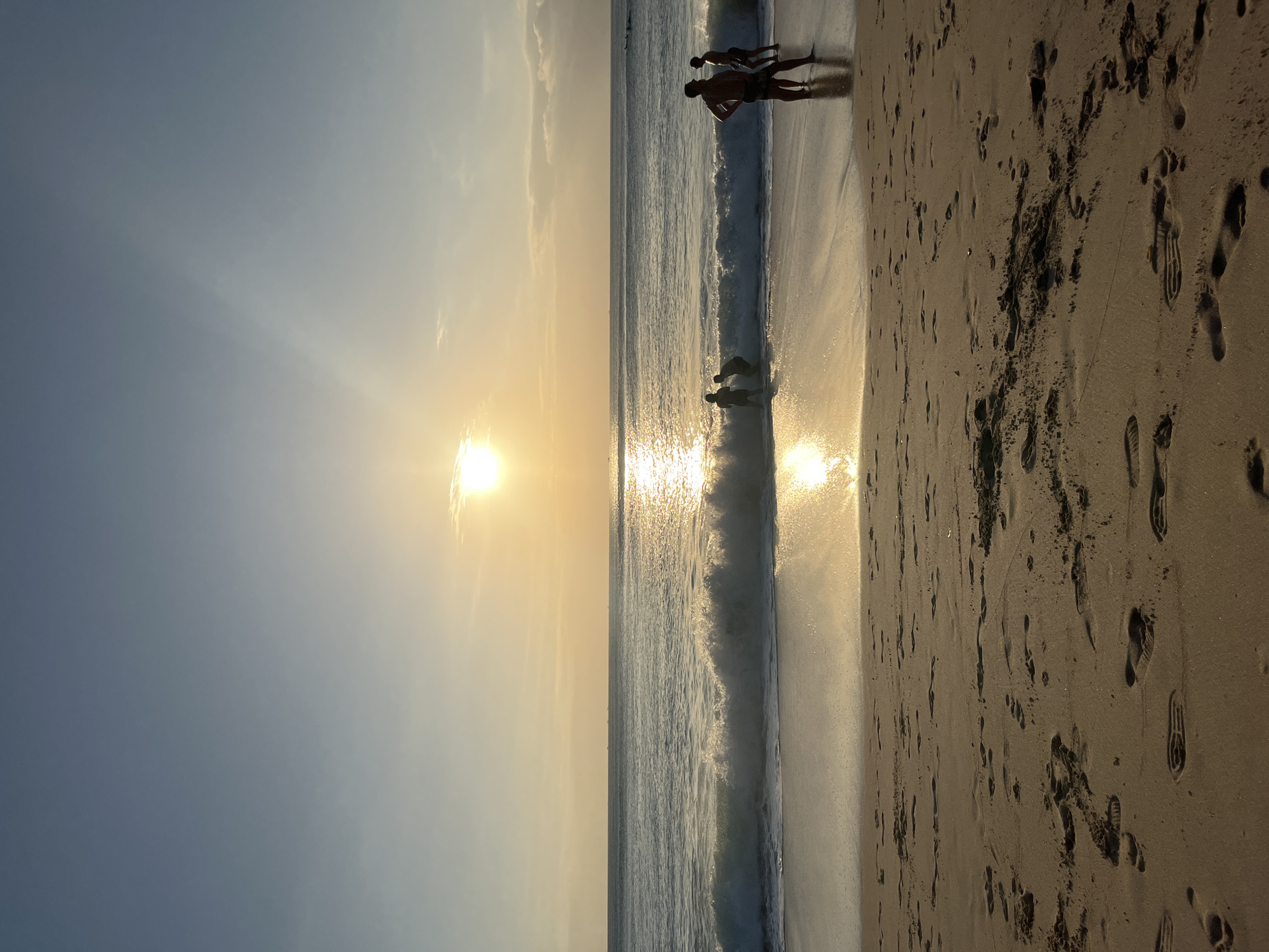 A sunset scene on a beach in Kuta Selatan, Indonesia, captures the golden light reflecting on the water and sand. The sun is low on the horizon, casting a warm glow over the scene. The ocean has gentle waves with white foam breaking on the shore. Several people are visible; two are standing in the shallow water, while another two are standing on the wet sand near the water's edge. The foreground is the sandy beach with numerous footprints. The sky is a gradient of blue and yellow, with some light clouds present. In the distance, several small boats are visible on the horizon line. The overall atmosphere is serene and tranquil.