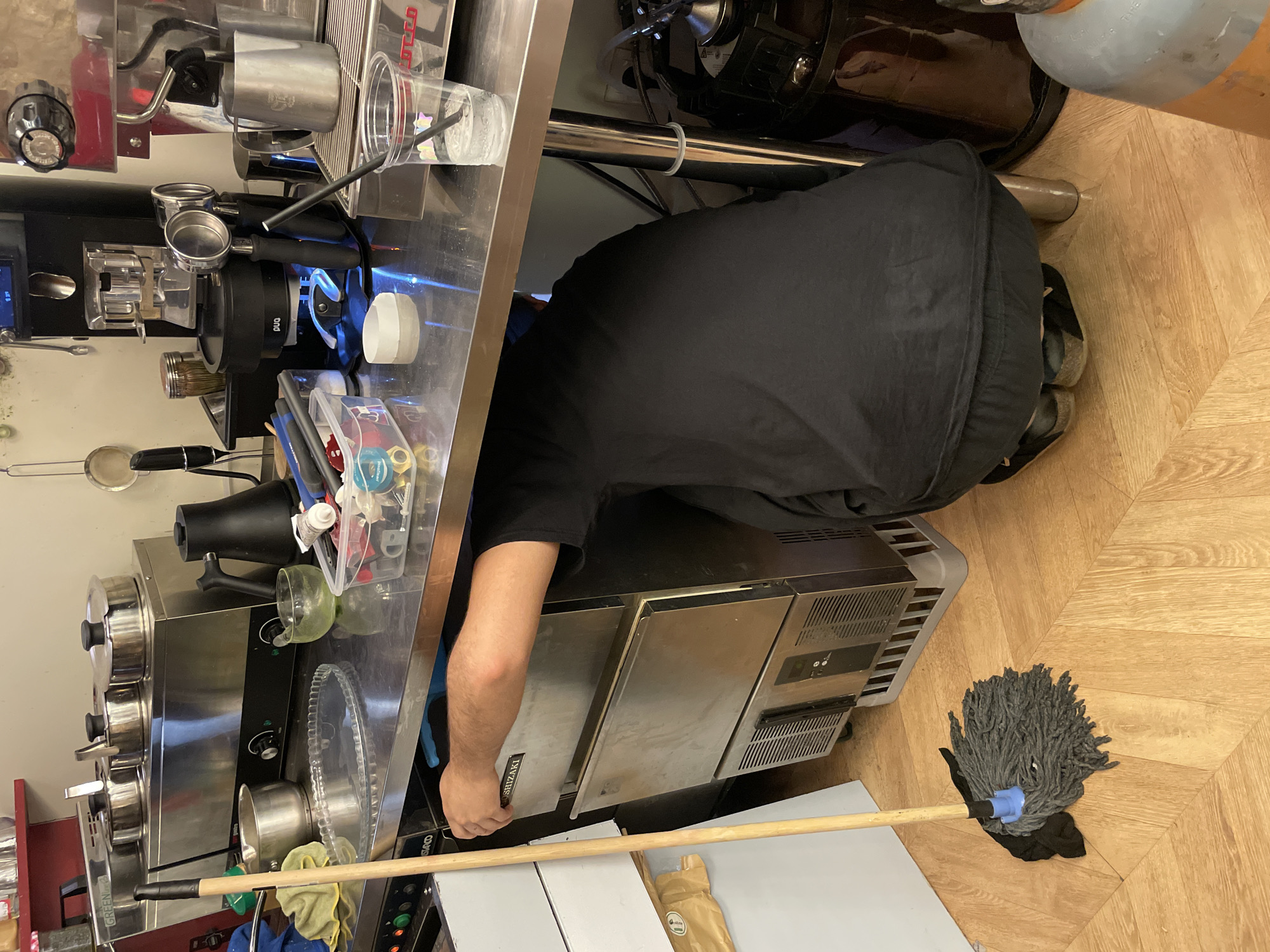 A person in a black t-shirt kneels in a commercial kitchen, apparently working on a Shizaki ice machine. A mop lies on the wood-patterned floor. An espresso machine and assorted containers are nearby. The location is not possible to identify.