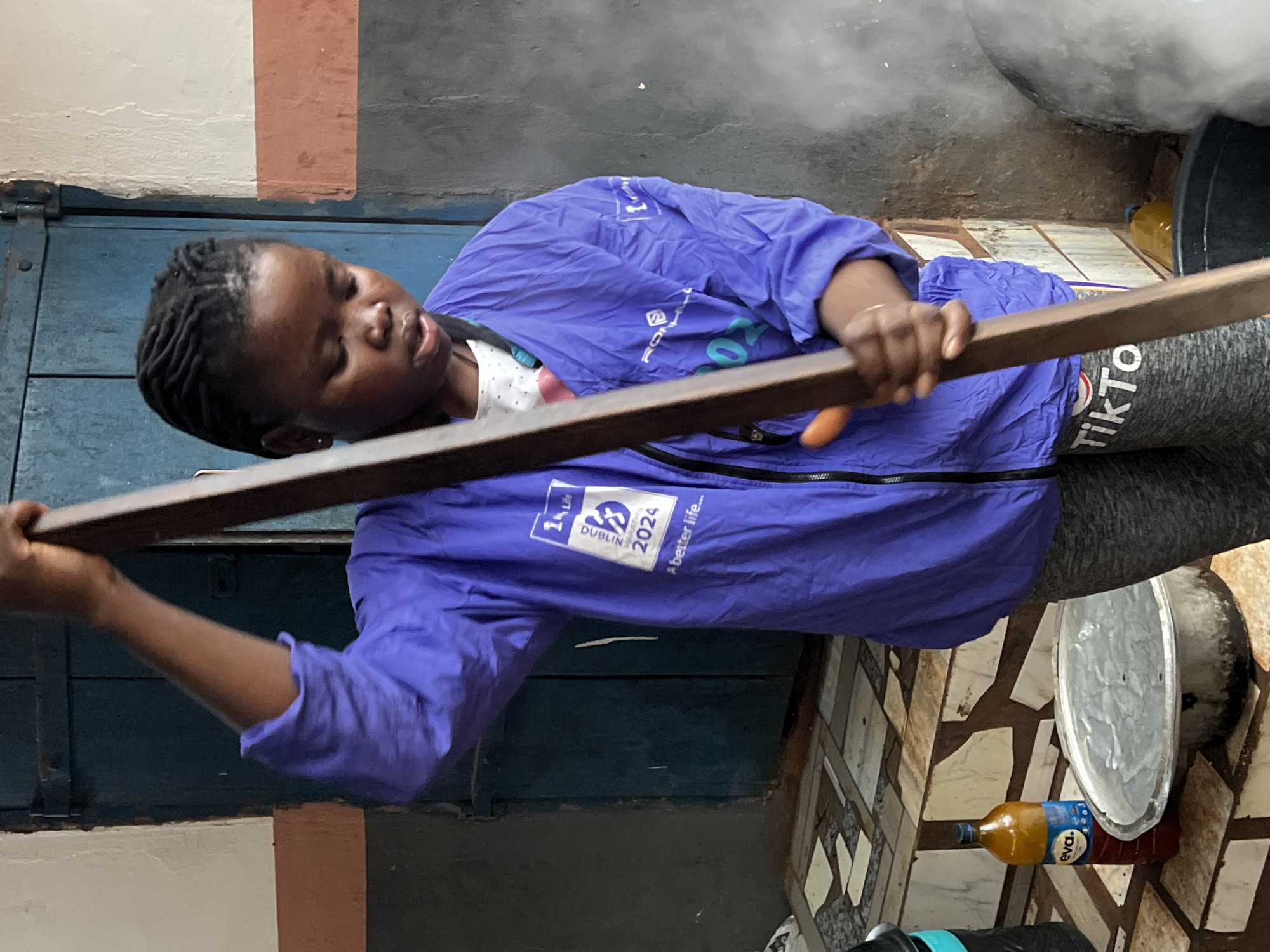 A young woman with dark skin and braided hair, wearing a vibrant purple jacket over a white shirt and grey leggings with "TikTok" printed on them, is stirring a large pot with a wooden spoon. The jacket has a logo on the left breast that reads "1Life Dublin Marathon 2024 A better life...". The background consists of a blue wooden door and a grey wall. In the foreground, to the left of the woman, there is a bottle of "Eva" cooking oil and a metal pot. The scene appears to be set in an outdoor or semi-outdoor location in Nkpor, Nigeria, possibly a kitchen or food preparation area, given the cooking implements and ingredients.