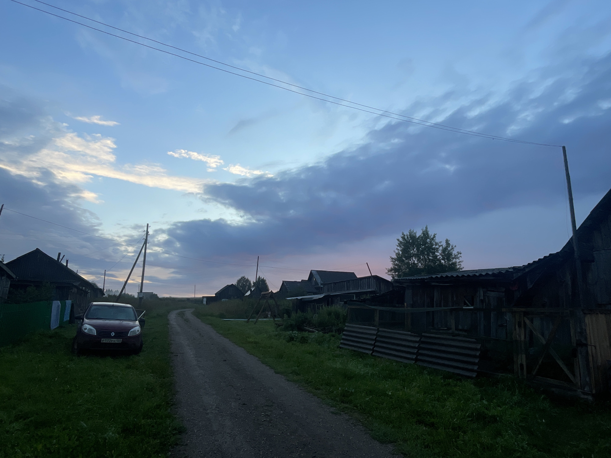 The image depicts a rural scene in Panteleevka, Russia, during twilight. A dirt road stretches into the distance, flanked by grassy verges and the silhouettes of wooden houses and outbuildings. The sky is a gradient of soft blues and purples, with scattered clouds catching the last rays of sunlight. A dark-colored car, possibly a Renault Sandero based on the visible badge, is parked on the left side of the road, its driver's side door ajar. Laundry, appearing to be sheets or towels, is draped over a fence near the car. The architecture of the buildings suggests a traditional village setting, with weathered wooden structures and corrugated metal roofing visible on some. Power lines crisscross the sky, supported by wooden poles, a common feature in rural Russian landscapes. The overall atmosphere is one of quiet, end-of-day calm in a remote settlement.