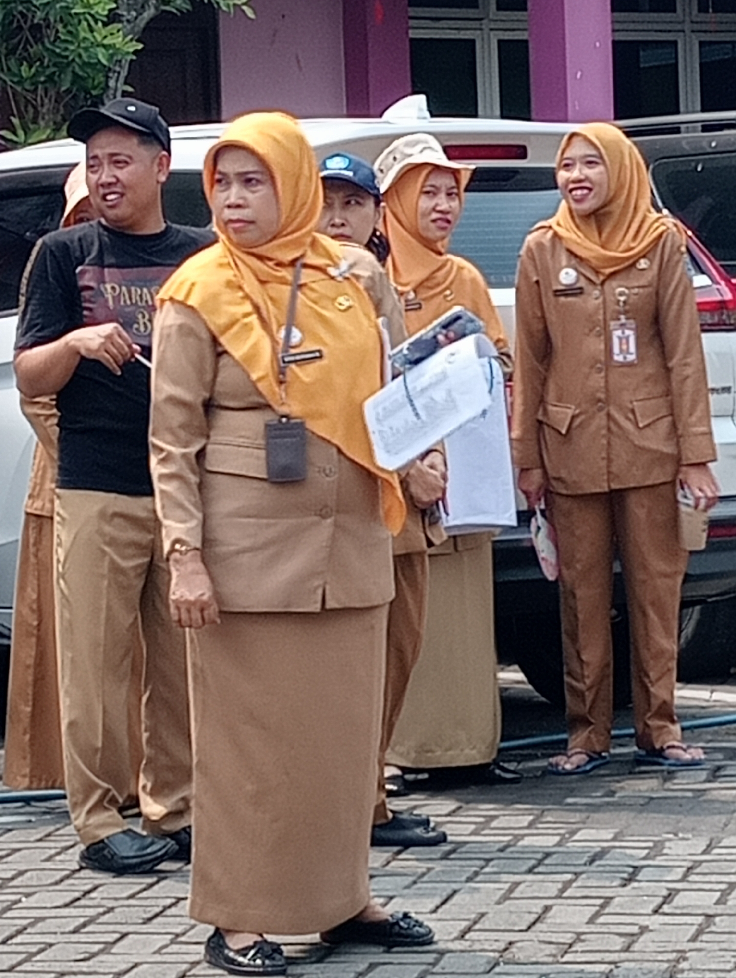 The image depicts a group of five individuals, three women and two men, standing outdoors on a paved surface. Two of the women are wearing tan-colored uniforms with headscarves, appearing to be government employees or civil servants, given the insignia on their uniforms. One woman in the foreground, also in a tan uniform and headscarf, is looking to her left and holding a white folder. Another woman in a similar uniform and headscarf, positioned further back, is smiling and looking to her right. A third woman, in a similar tan uniform and headscarf, is in the far background, looking to her right.
The man on the left, wearing a black t-shirt with a graphic print and a black baseball cap, is looking to his left with an open mouth. The second man, in the far background, is partially obscured and wearing a blue cap. The setting appears to be the exterior of a building with a pink facade visible in the background. The presence of vehicles suggests an outdoor parking or gathering area. While the specific location in Demak, Indonesia, is not explicitly identifiable from the visual cues alone, the attire of the women suggests a formal or official context. The overall scene captures a moment of interaction or activity among the group.