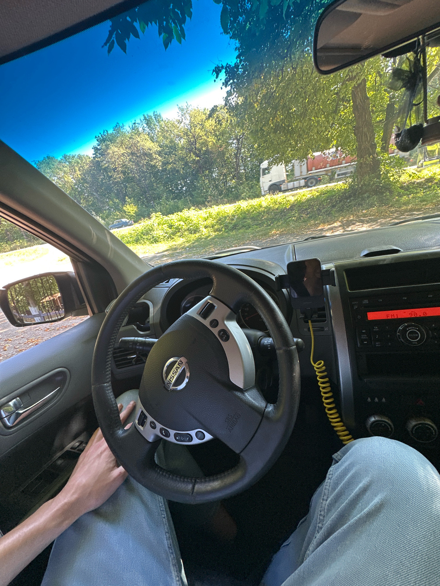The image shows the interior of a car, likely a Nissan, from the perspective of the driver. The steering wheel with the Nissan logo is prominent in the foreground. A person's hands are visible on the steering wheel and their legs are covered in light blue jeans. A smartphone is mounted on the dashboard using a black holder, connected to a coiled yellow charging cable. The car's radio display shows "FM1 90.0". Outside the car, through the windshield and side windows, a wooded area with green trees and sunlight filtering through is visible. In the background, a white truck with a red tanker trailer is parked on a dirt road. The location is identified as Shatava, Ukraine.
