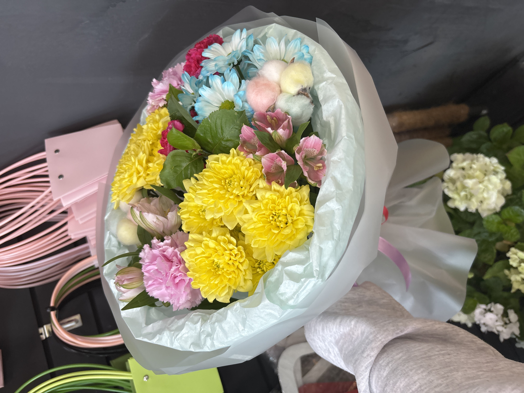 An overhead medium shot captures a bouquet of vibrant flowers wrapped in light blue and white paper, held by a person wearing a grey sweater. The bouquet is densely packed with a variety of blooms, including bright yellow chrysanthemums, pink carnations, light pink alstroemerias, and small blue and white daisies. Interspersed are fluffy cotton-like accents in pastel shades of pink, yellow, and blue. In the background, out-of-focus pink and green shopping bags hang on a black surface, along with some looped pink and green material. The lighting is soft and natural, casting subtle shadows and highlighting the textures of the flowers and wrapping paper. The scene appears to be indoors, possibly in a shop or market.