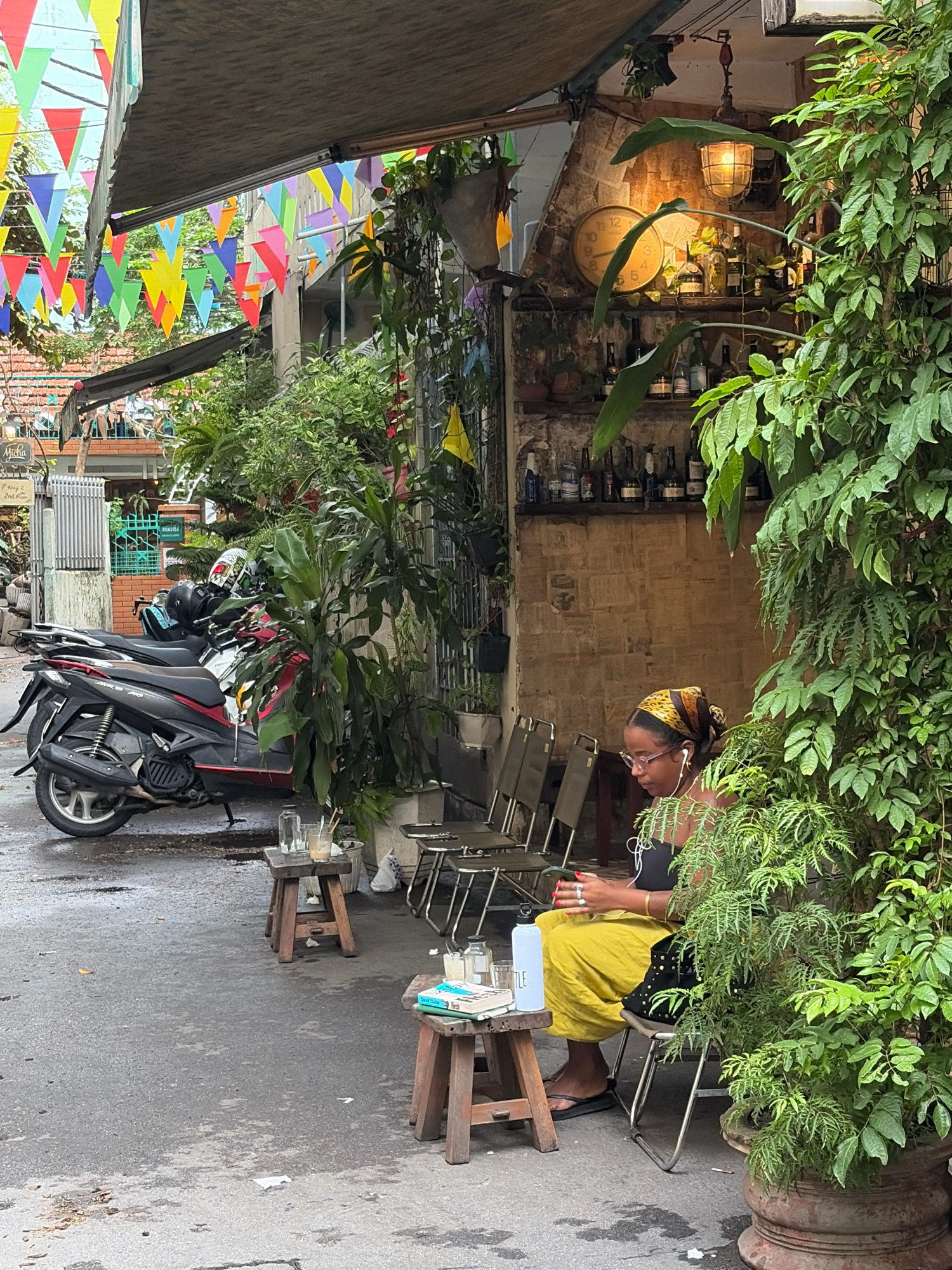 A woman sits on a metal chair at an outdoor cafe in Da Nang, Vietnam, wearing a yellow skirt, a black top, and a patterned headscarf. She has earbuds in and is looking at her phone. In front of her, on a small wooden stool, is a light blue water bottle and a book. To her right, a large potted plant with green leaves fills the frame. In the background, there are shelves with bottles, a clock, and a light fixture. Several motorcycles are parked to the left of the cafe. Colorful triangular flags are strung above the cafe's awning. The ground is wet, suggesting it may have recently rained.
